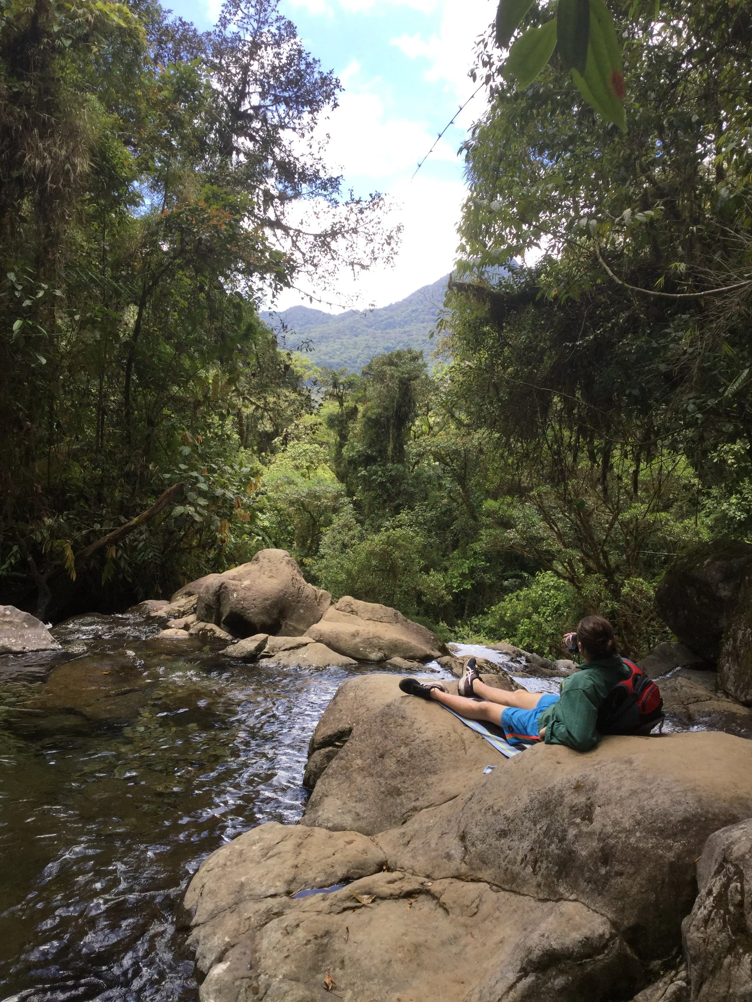 A hiker resting on rocks beside flowing water while looking across the cloud forest and green mountains near Boquete, Panama.