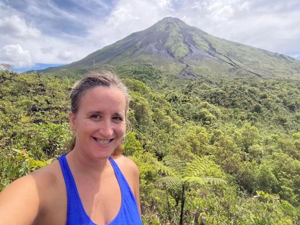 View of Arenal Volcano rising above the Costa Rican rainforest, with a smiling traveller standing in the foreground at a scenic viewpoint.