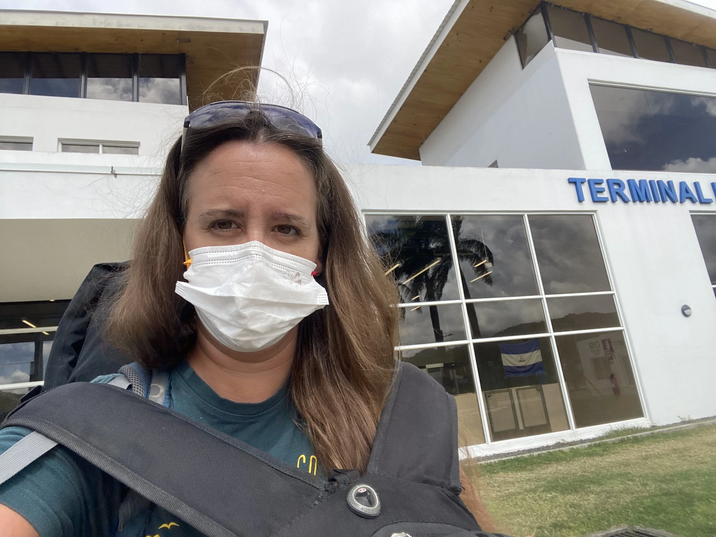 Female backpacker wearing a face mask and carrying a large travel backpack outside a terminal building in Central America.