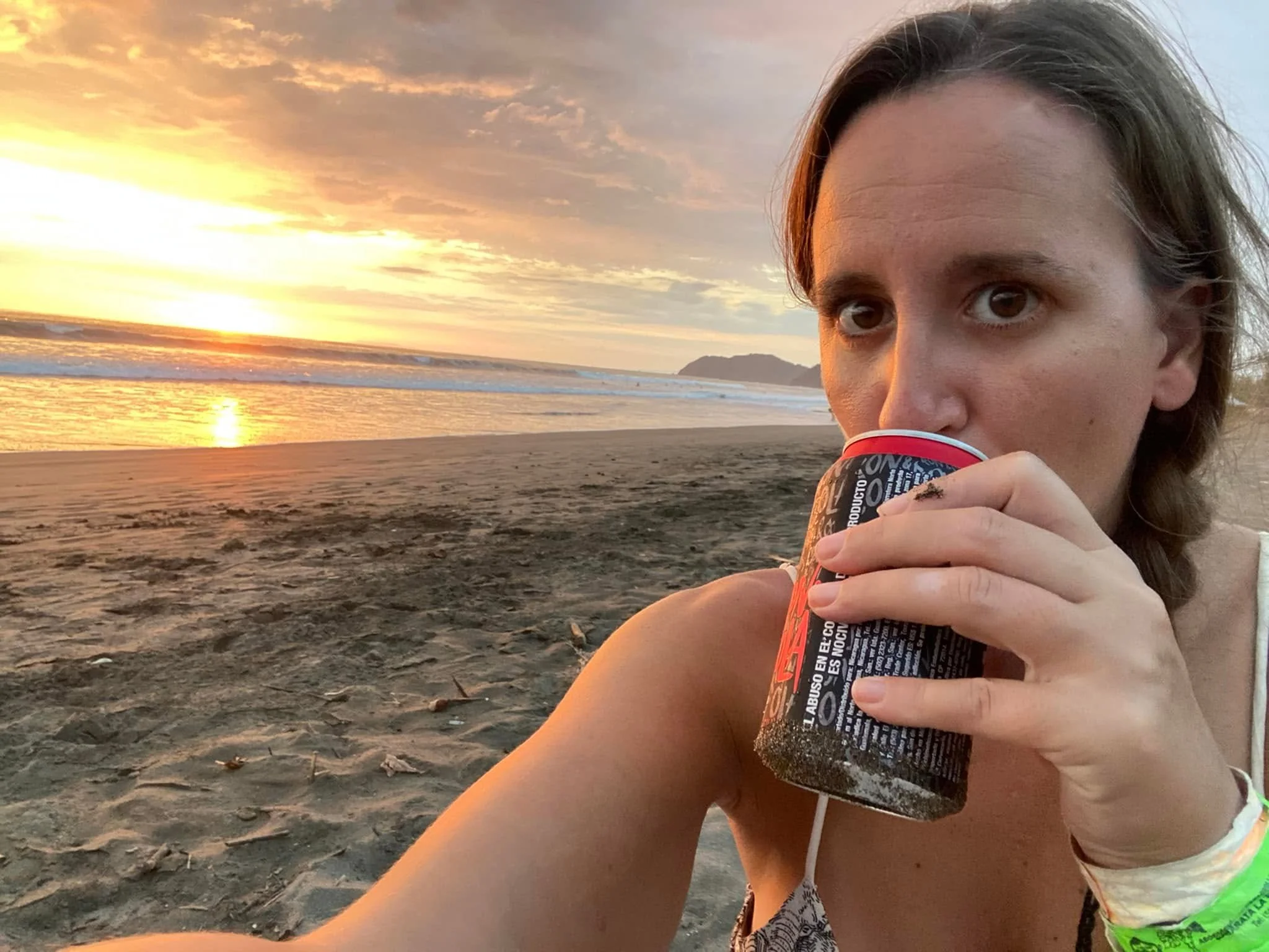A person sitting on Jacó Beach at sunset, drinking a canned rum and coke with the Pacific Ocean and golden sky in the background.