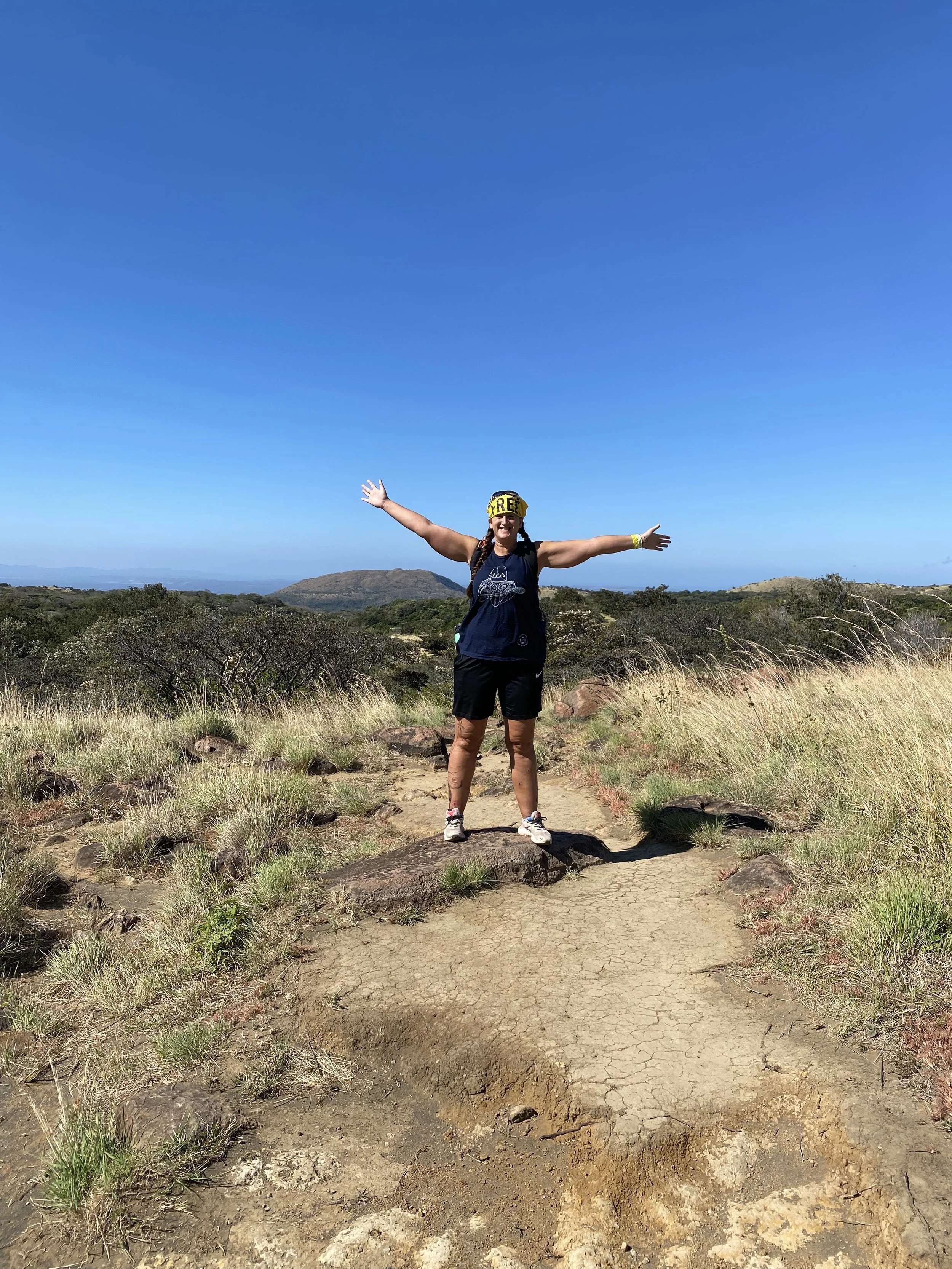 Backpacker standing on a hiking trail in Rincón de la Vieja National Park with arms outstretched and rolling hills in the background.
