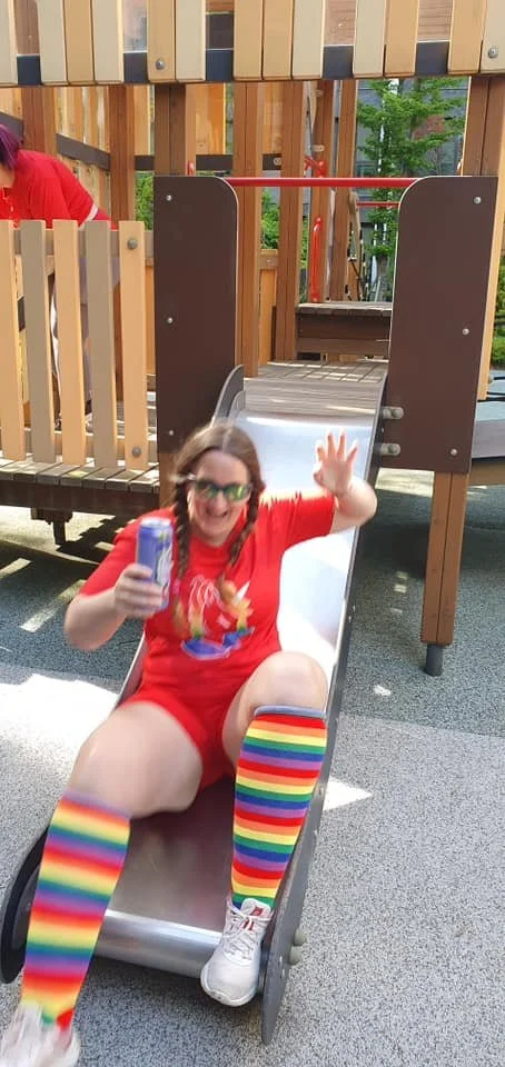 A woman wearing rainbow socks and a Pride shirt while sliding down a playground slide.