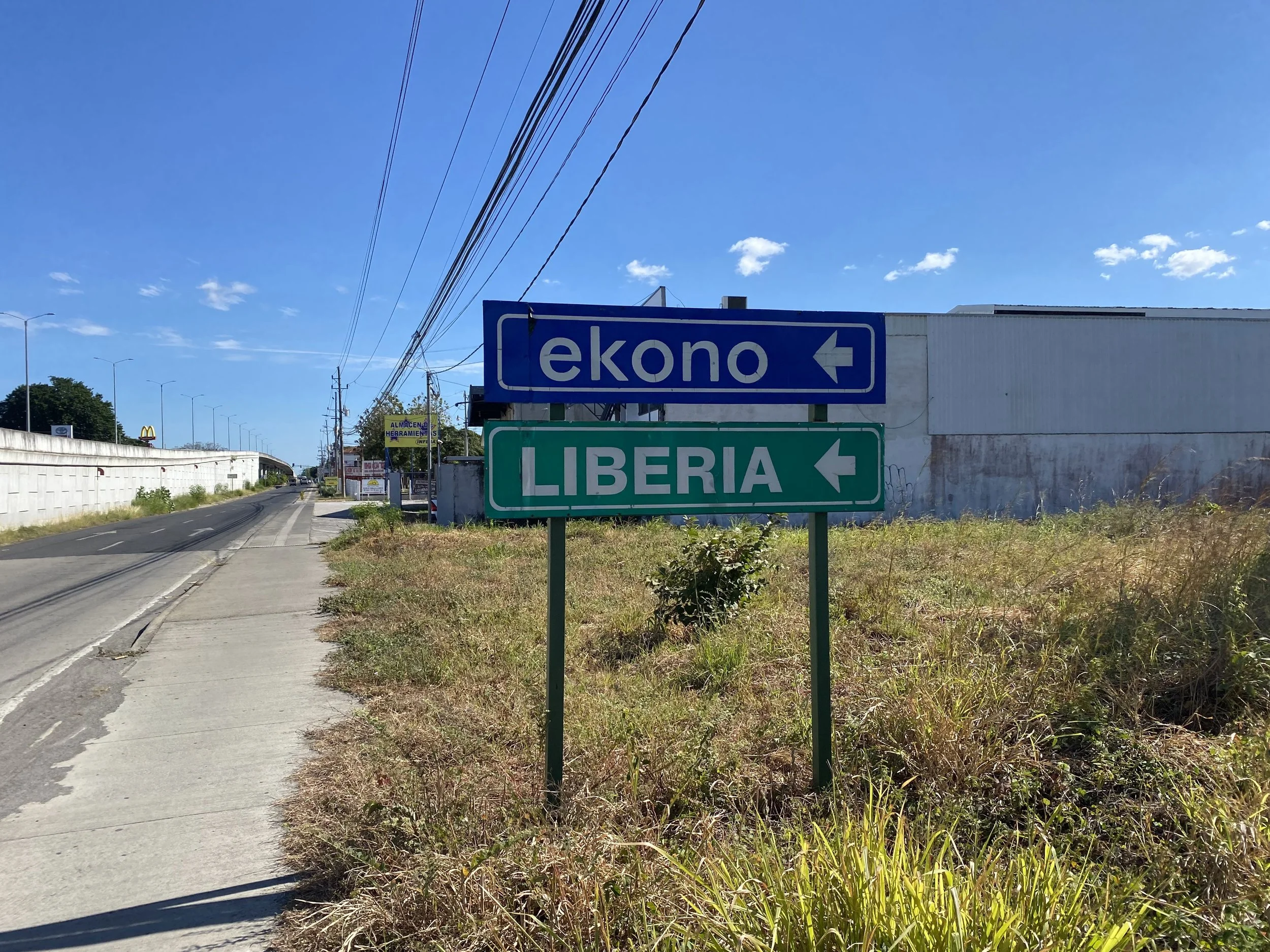 Roadside sign pointing toward Liberia, Costa Rica, beside a sidewalk where the backpacker began walking after arriving by shuttle.