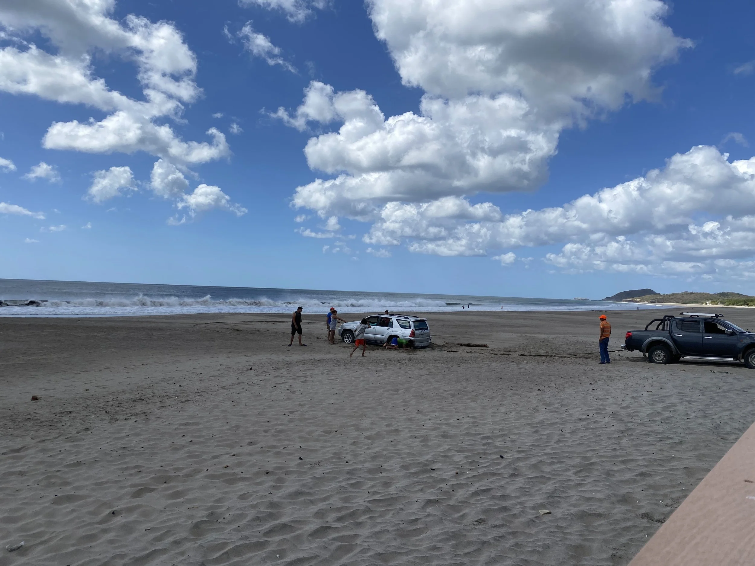 A vehicle stuck in the sand on Popoyo Beach in Nicaragua while several people work together to free it, with the Pacific Ocean and blue skies in the background.