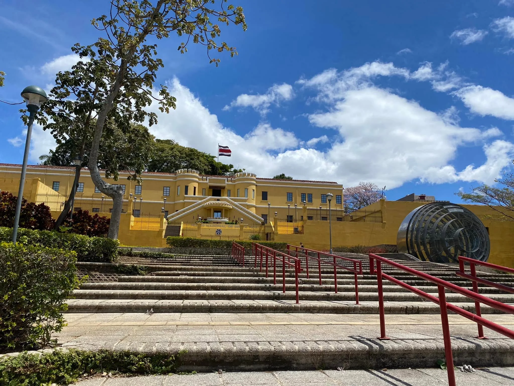 The yellow exterior of the National Museum of Costa Rica above wide stone steps and red railings, with a Costa Rican flag flying under a bright blue sky.