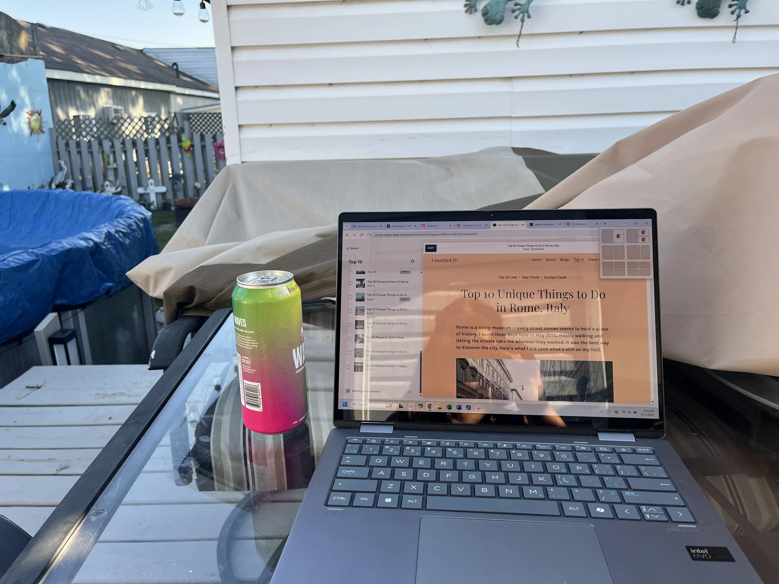 A laptop displaying the Unsettled JD travel website on an outdoor patio table beside a canned drink, showing the behind-the-scenes process of writing and publishing travel articles.