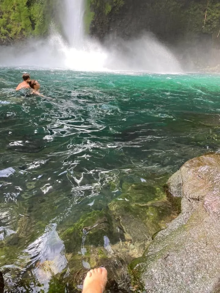 A foot dipping into the clear pool at La Fortuna Waterfall in Costa Rica, showing the cold water and rocky edge.