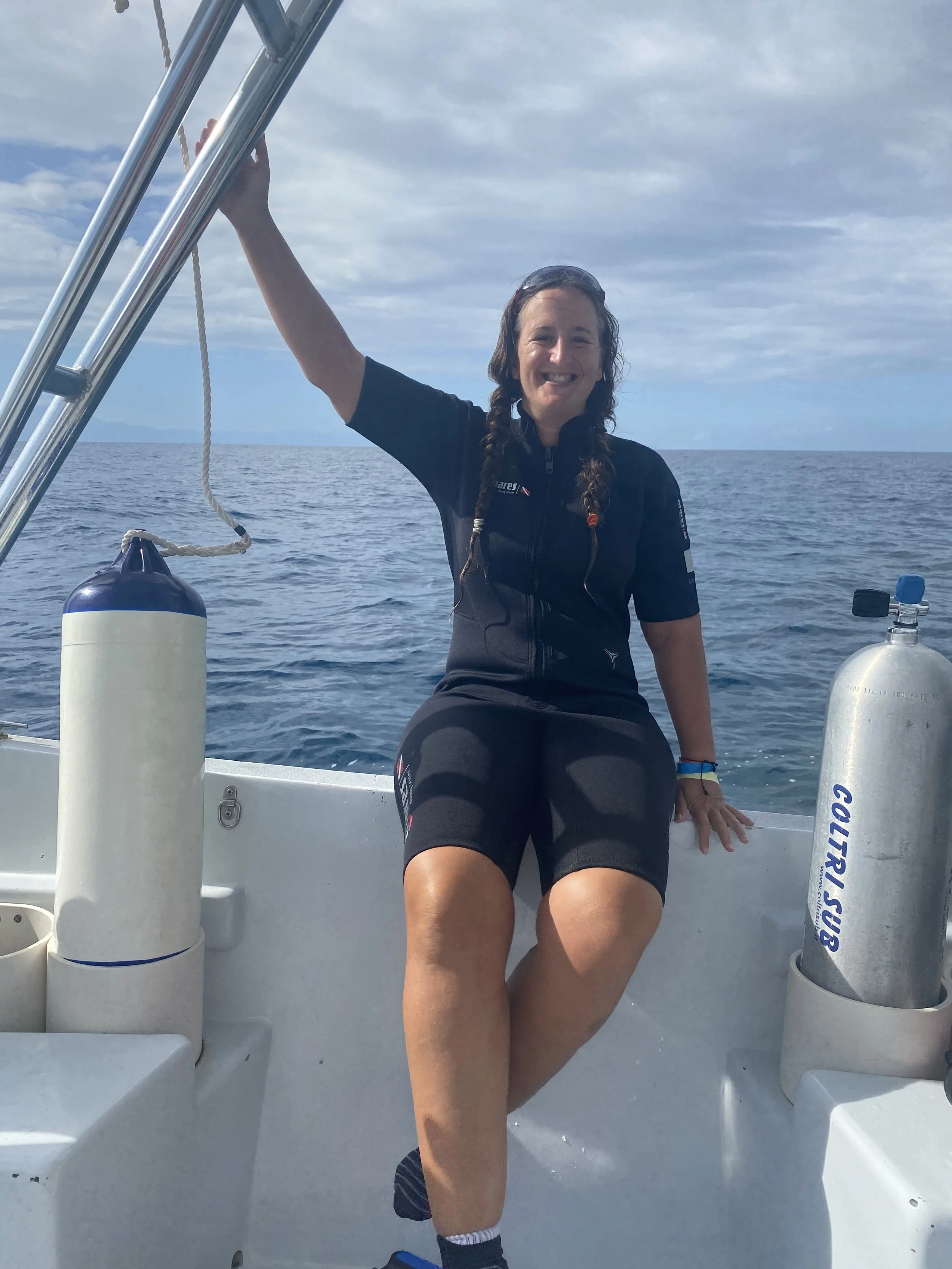 Woman in a wetsuit sitting on the edge of a dive boat in Utila with scuba tanks and open ocean behind her.