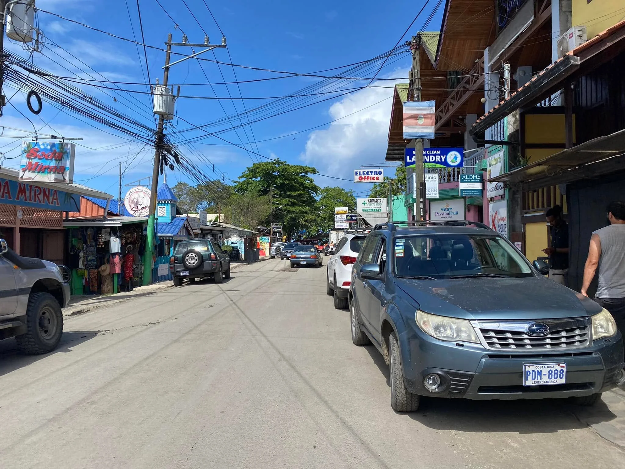 Busy street in Puerto Viejo, Costa Rica, lined with colourful shops, restaurants, parked cars, tropical trees, and overhead power lines beneath a blue sky.