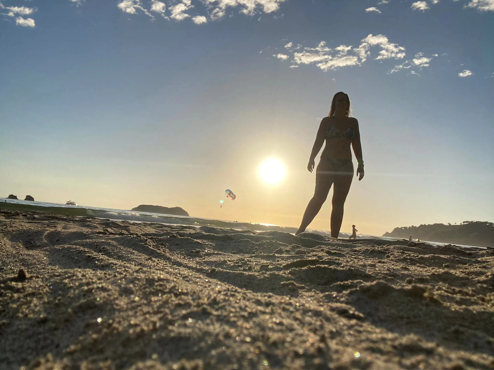 Silhouette of a woman standing on a sandy beach at sunset in Puerto Viejo, Costa Rica, with the Caribbean Sea and a parasail visible in the background.