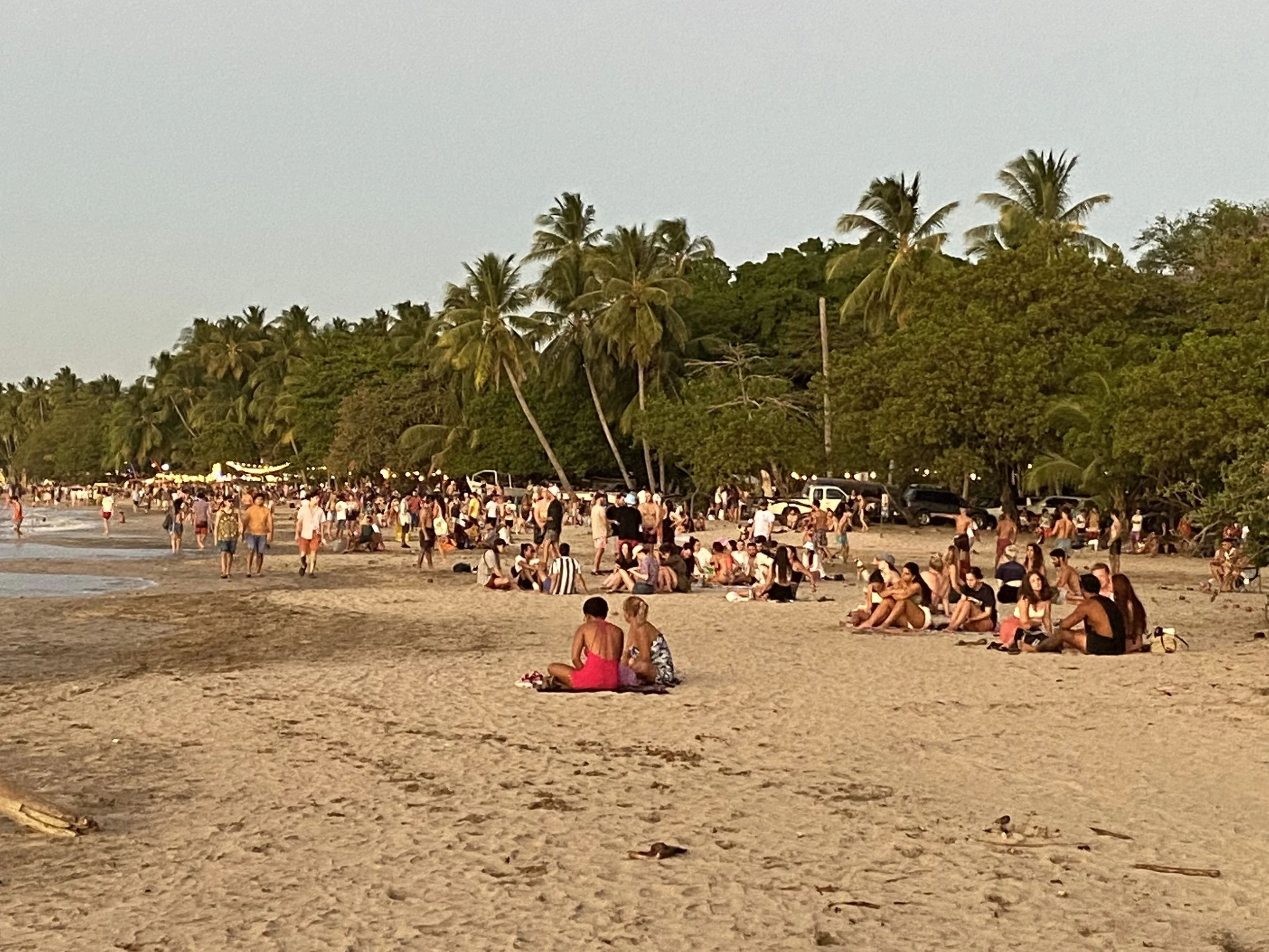 Crowds of people relaxing and walking along Tamarindo Beach in Costa Rica during sunset, with palm trees, the Pacific Ocean, and a lively beach atmosphere.