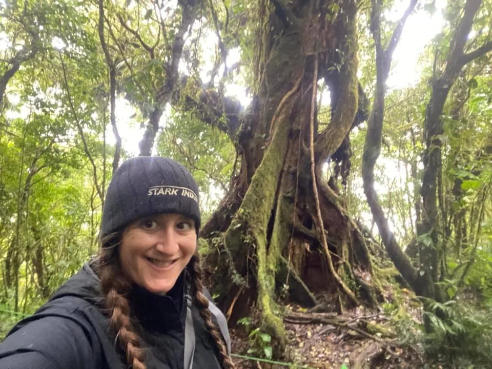 Woman wearing a winter hat and jacket standing beside a giant moss-covered tree in the Monteverde Cloud Forest, Costa Rica.
