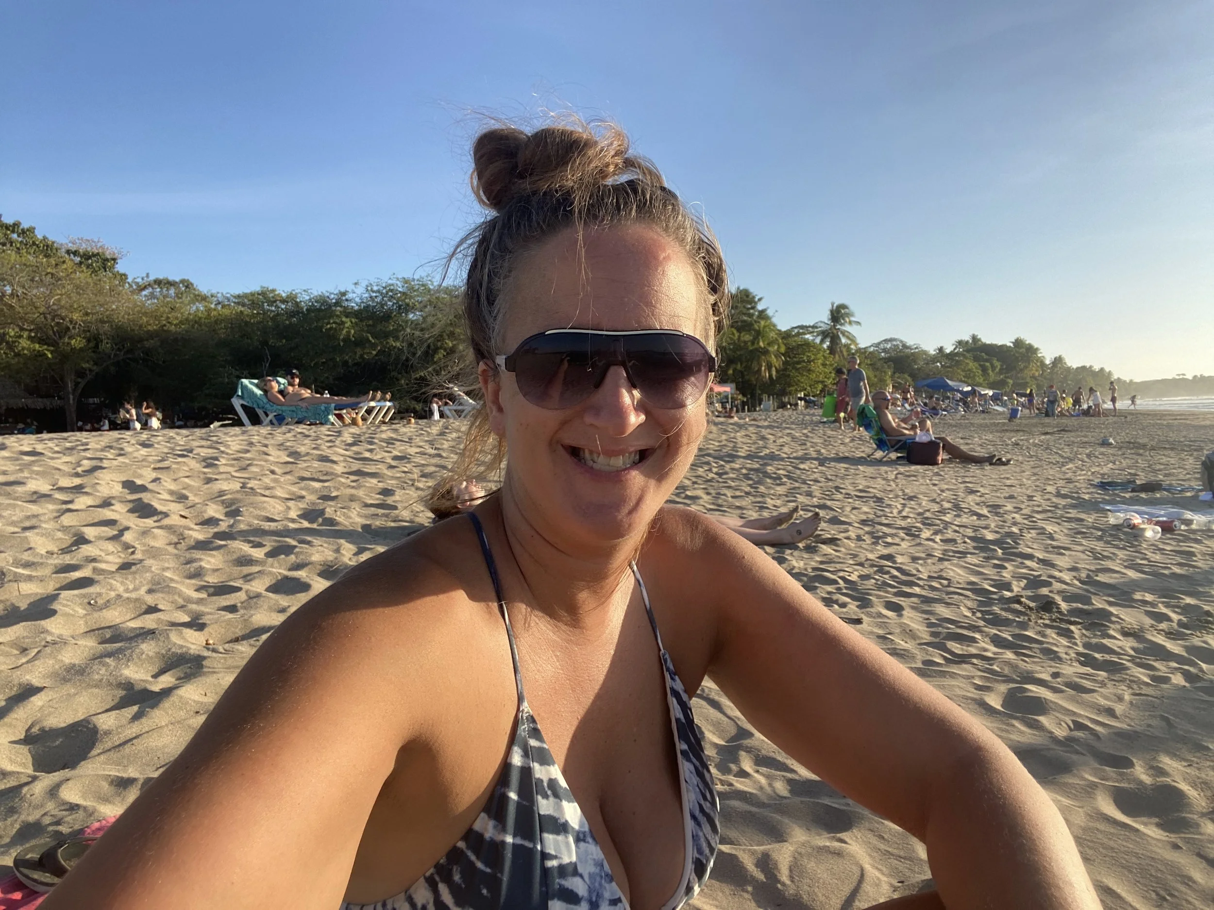 A woman sitting on the sandy beach in Tamarindo, Costa Rica, smiling at the camera with beachgoers, palm trees, and the Pacific coastline in the background.