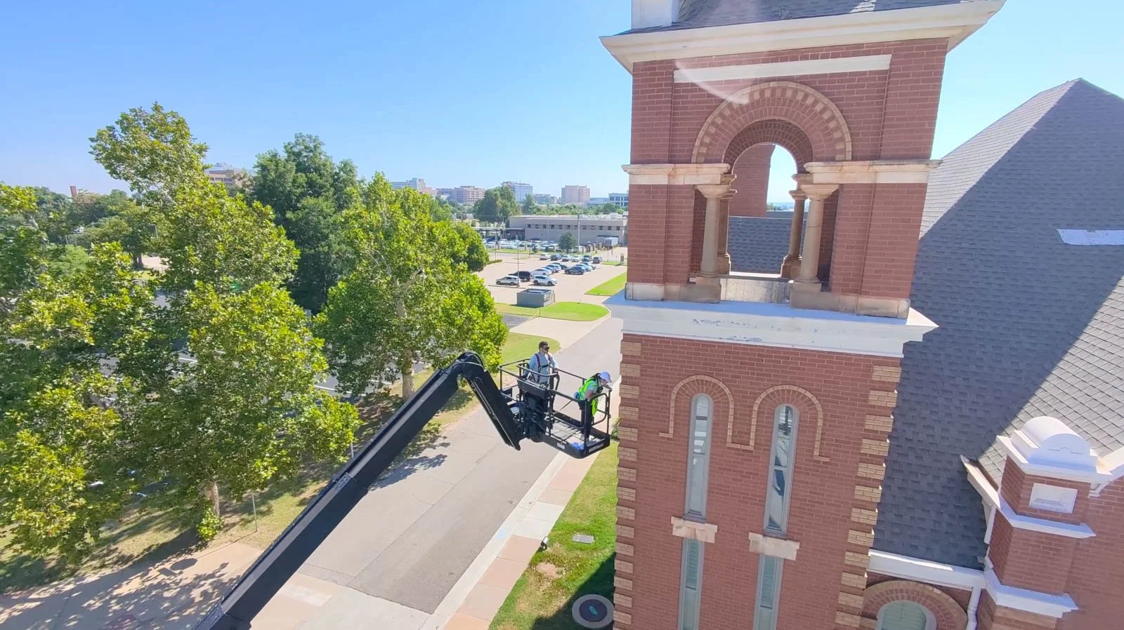 Two workers on a lift inspecting a clock tower on a red brick building with a parking lot and trees in the background.
