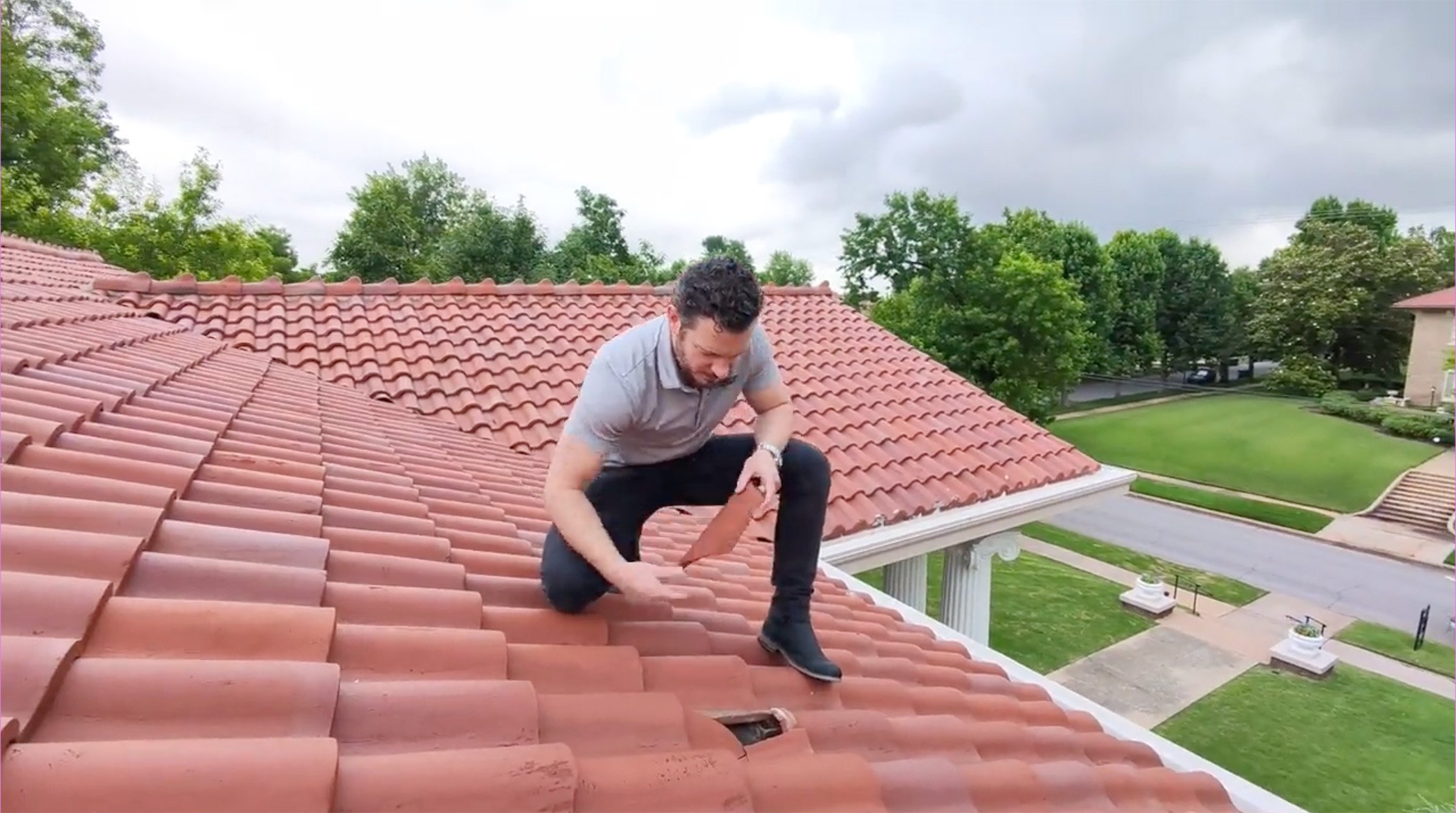 Man inspecting or repairing red clay tiled roof on a high building, with trees and a park visible in the background.
