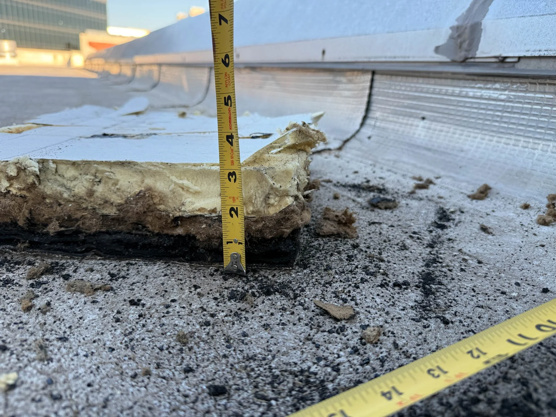 Close-up of a damaged hood insulation layer and exterior siding of a building, with measuring tapes showing height of approximately 6 inches. Debris and dirt are on the ground.