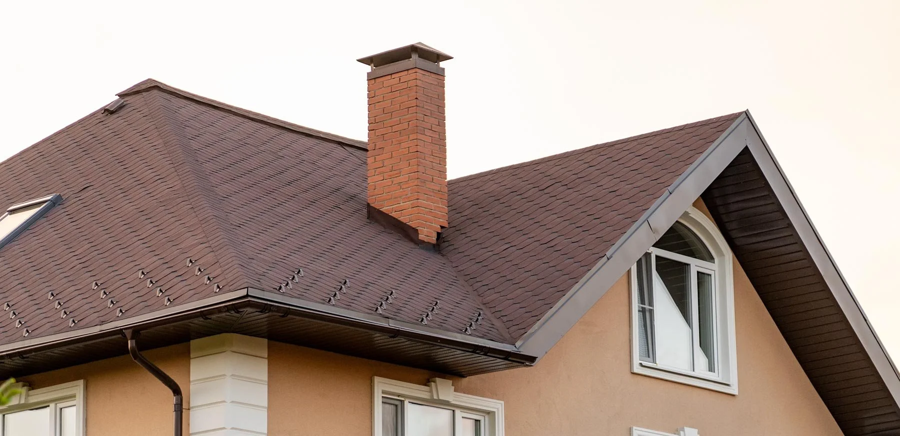 Close-up of a house's roof with brown shingles, a red brick chimney, and a large arched window in the attic.