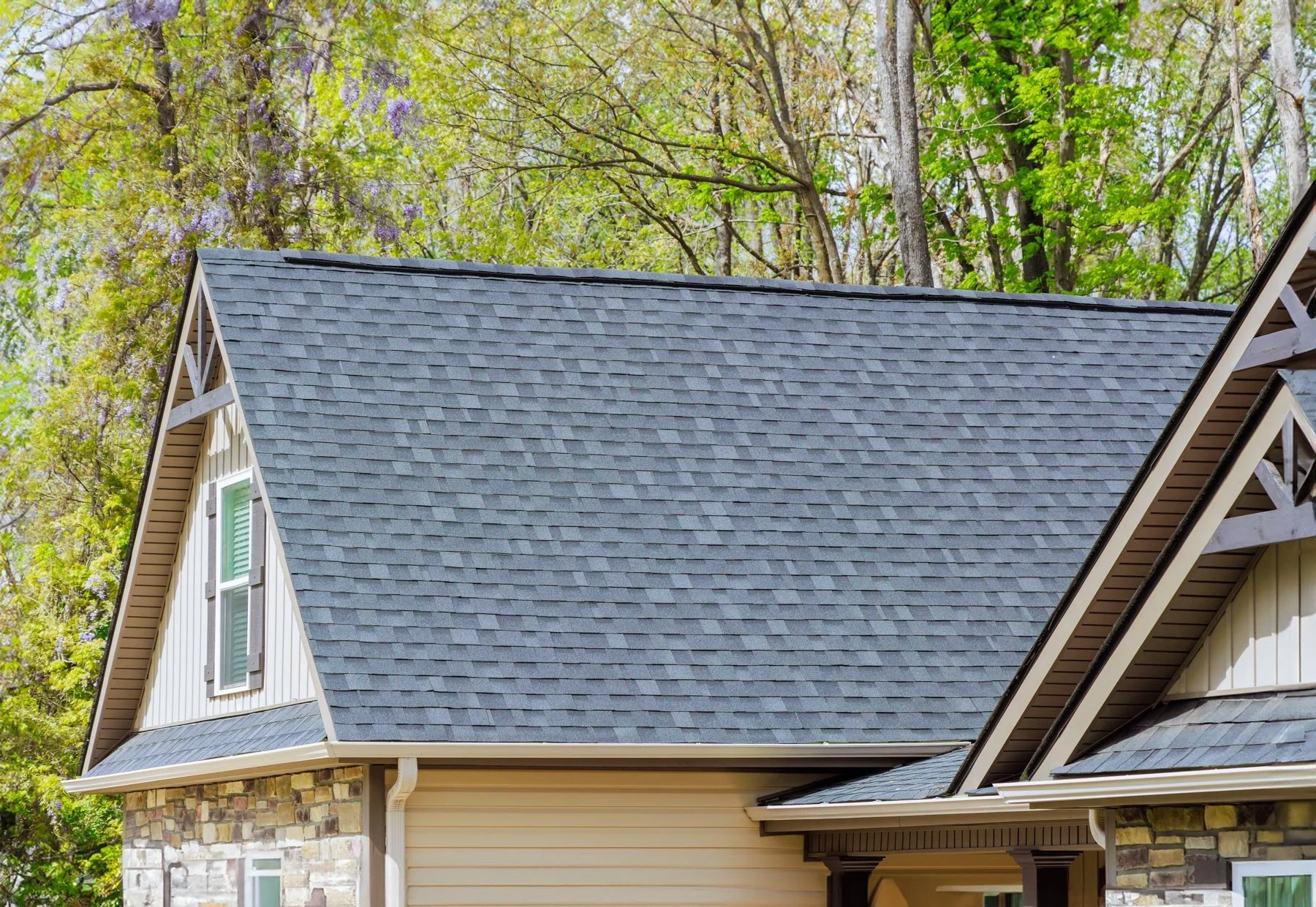 Close-up of a house roof with asphalt shingles, beige siding, and stone accents, with a background of leafy trees.