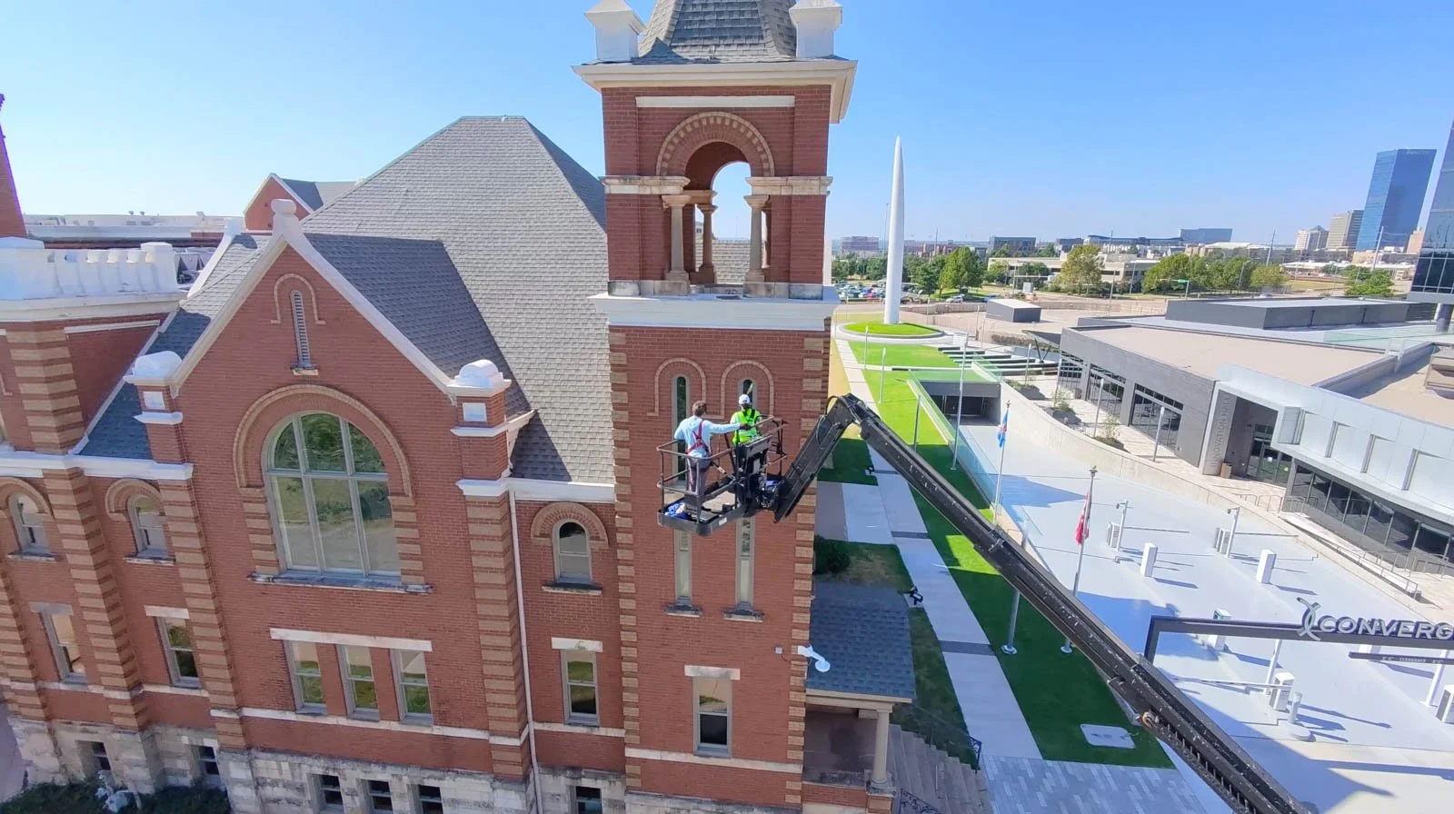 Two workers in safety gear on a cherry picker working on a red brick building with a tower and clock face, in an urban area with modern buildings, a green lawn, and a walkway.