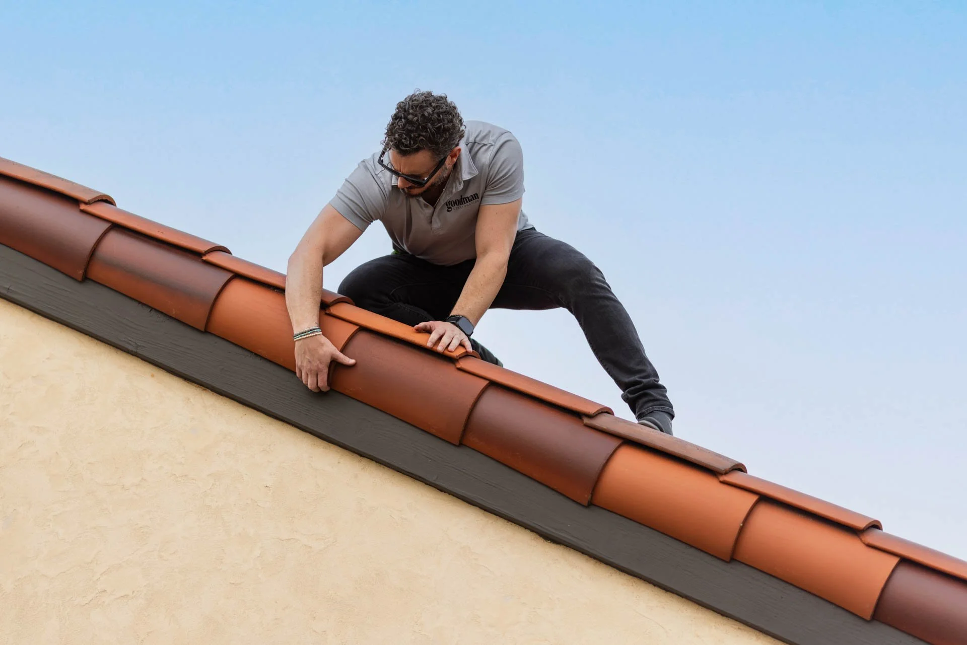 A man wearing a gray shirt and black pants is working on the roof of a building, installing or repairing red clay tiles.