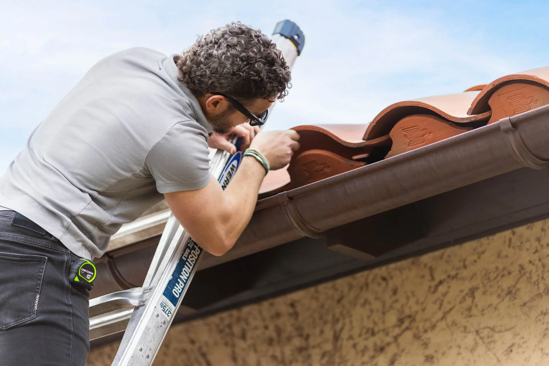 A man inspecting a roof with clay tiles, standing on a ladder.