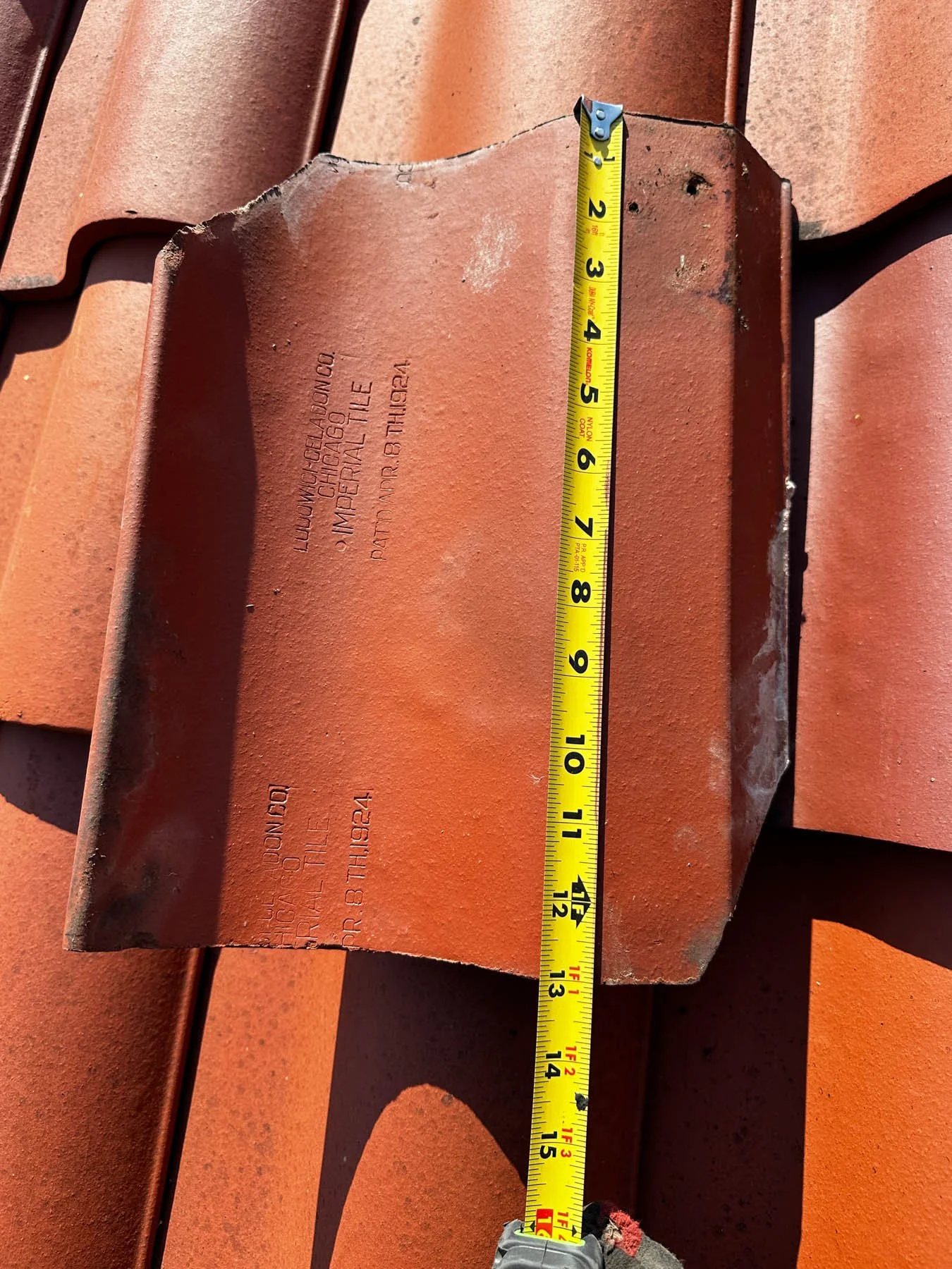 A measuring tape shows approximately 13 inches on a rusty orange roof tile with stamped markings.