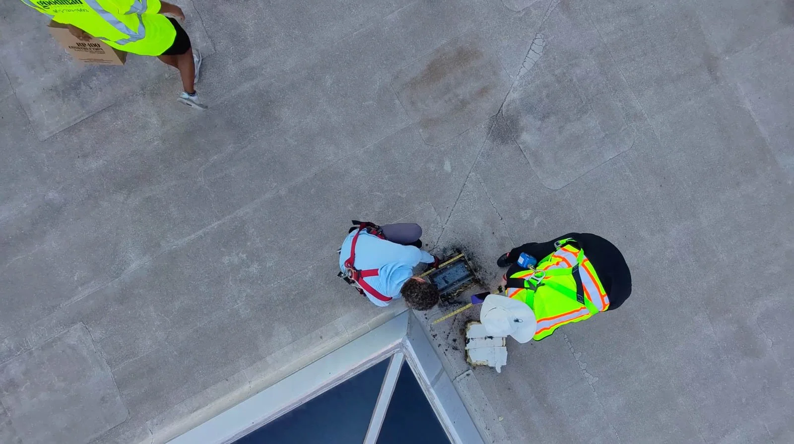 Two workers in safety vests working on a maintenance or repair task. One worker is kneeling down, using tools, and the other is standing nearby, observing or assisting.