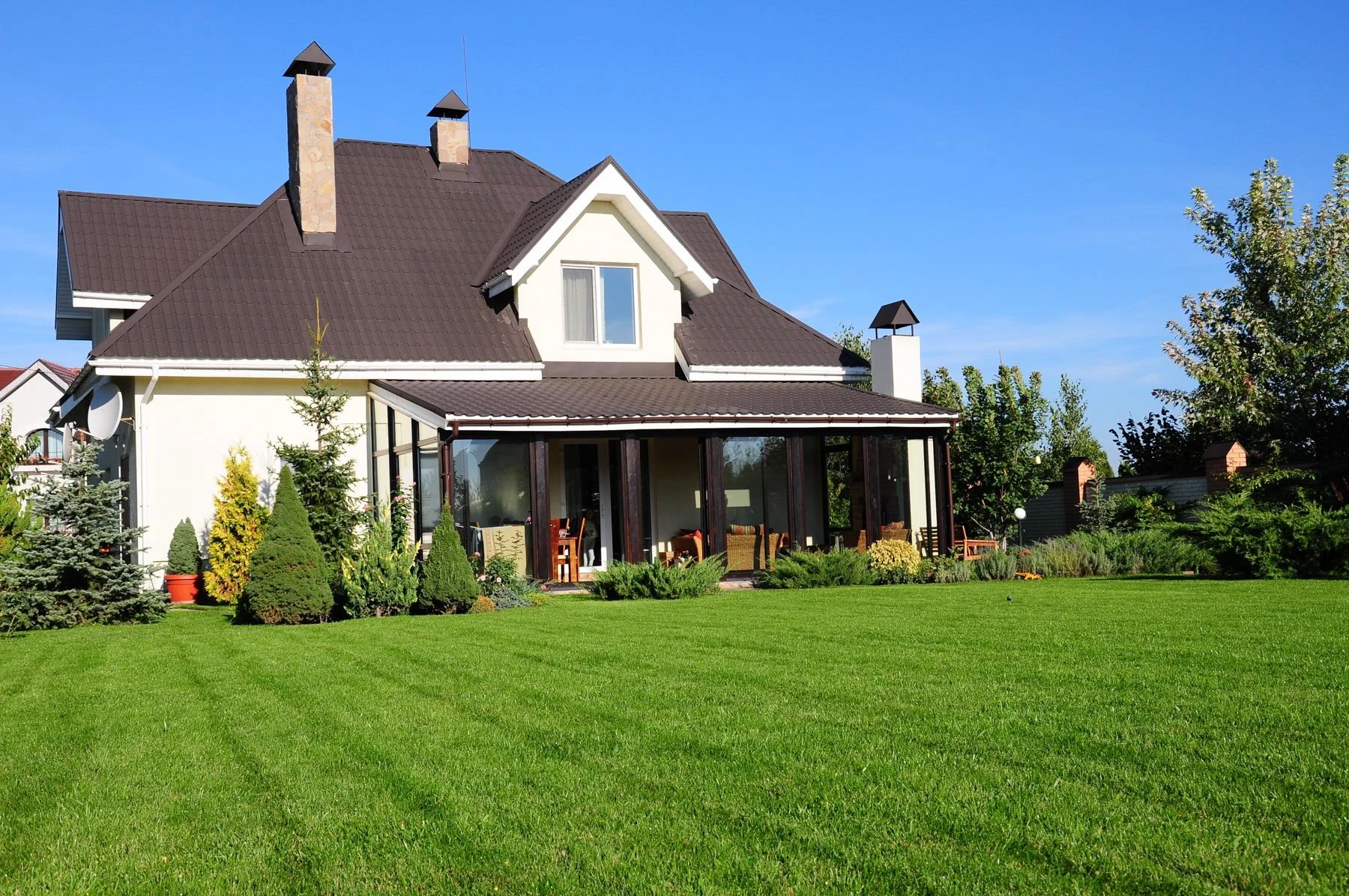 A large house with white walls, a dark-colored roof, and a glass-enclosed porch, surrounded by well-maintained green lawn and various trees and shrubs, under a clear blue sky.