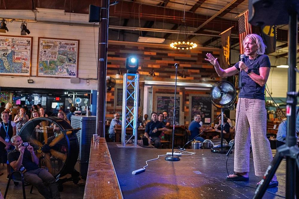 Kate Williams stands on a stage with a microphone, speaking to an audience in a restaurant or bar with wooden walls. The audience is seated at tables, watching her.