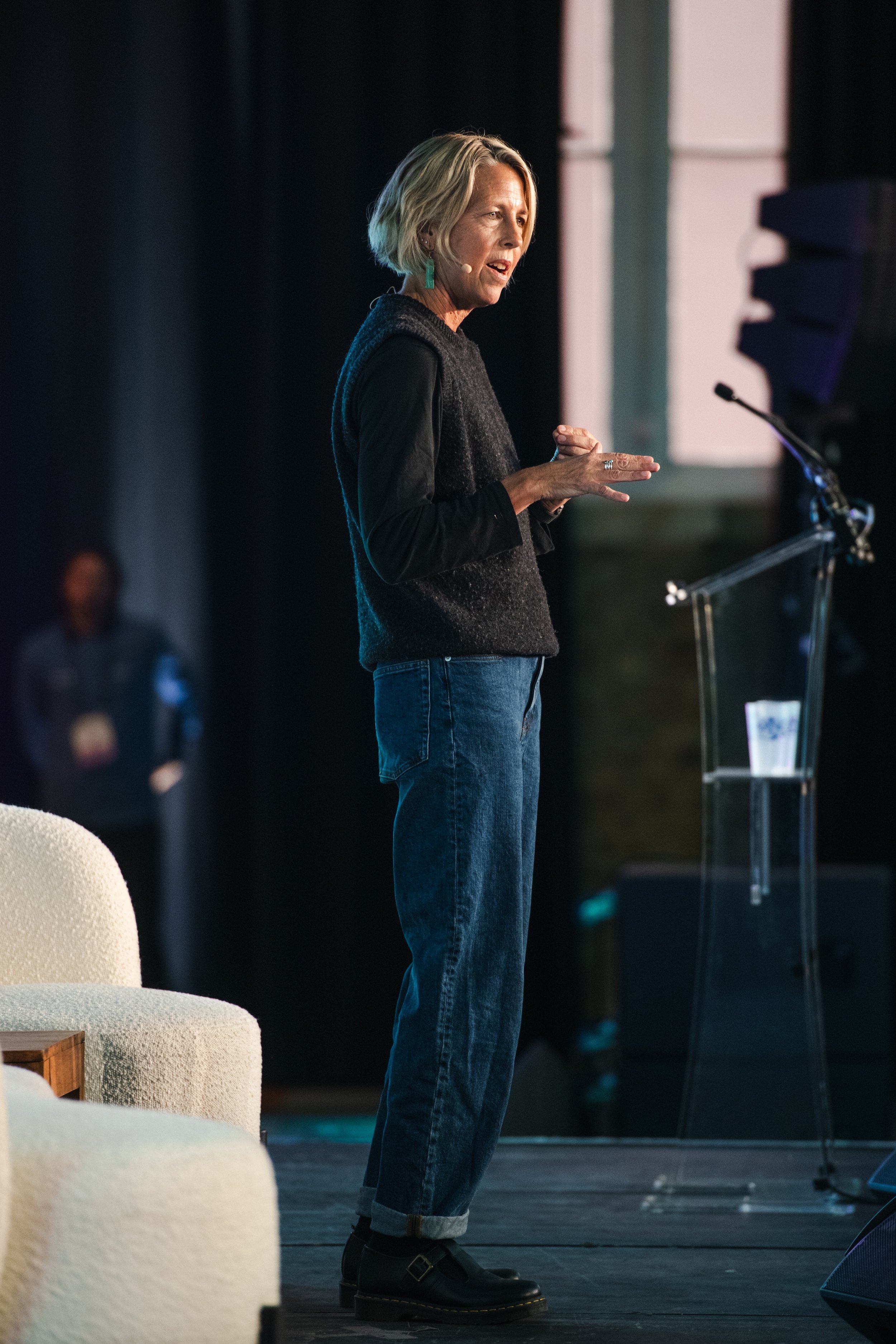 Kate Williams speaking at a podium on stage, wearing a black sweater, jeans, and black shoes, with a headset microphone.