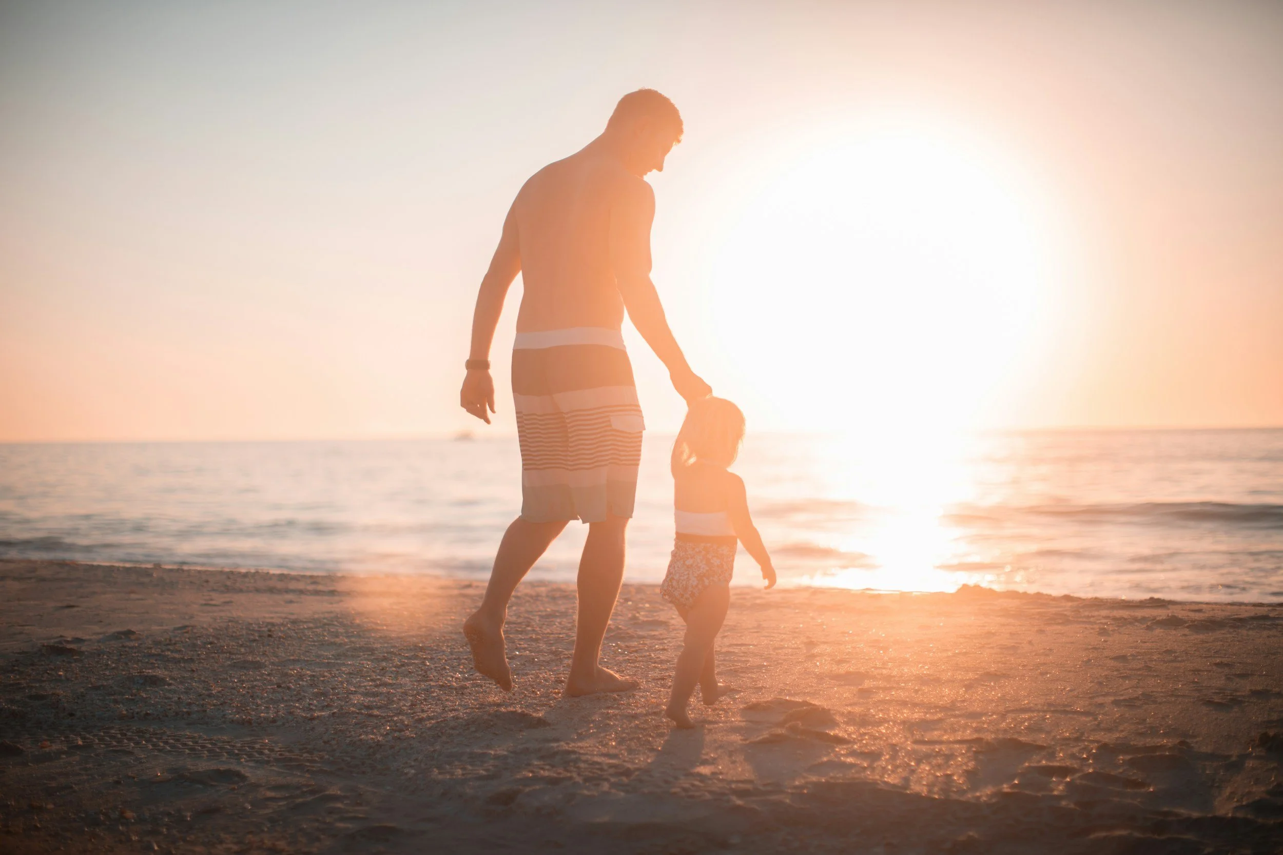 A man and a young girl walking barefoot on the beach at sunset, holding hands.
