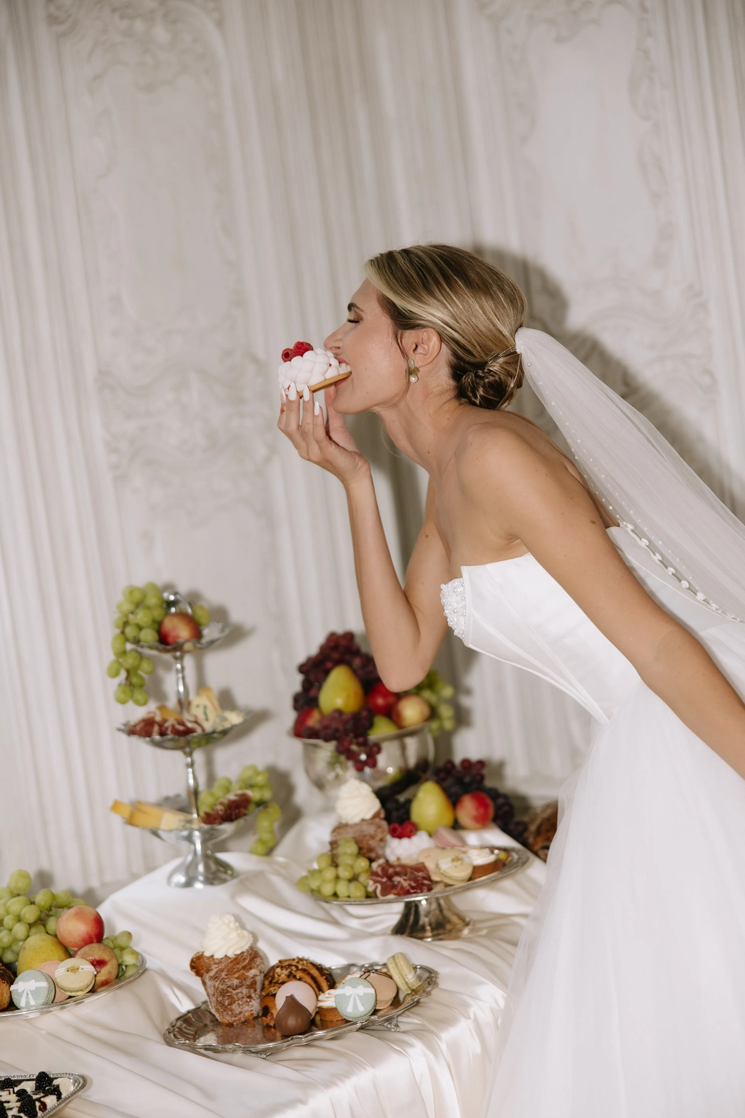 Bride in wedding dress with veil enjoying a slice of cake at a dessert table with fruit and pastries.