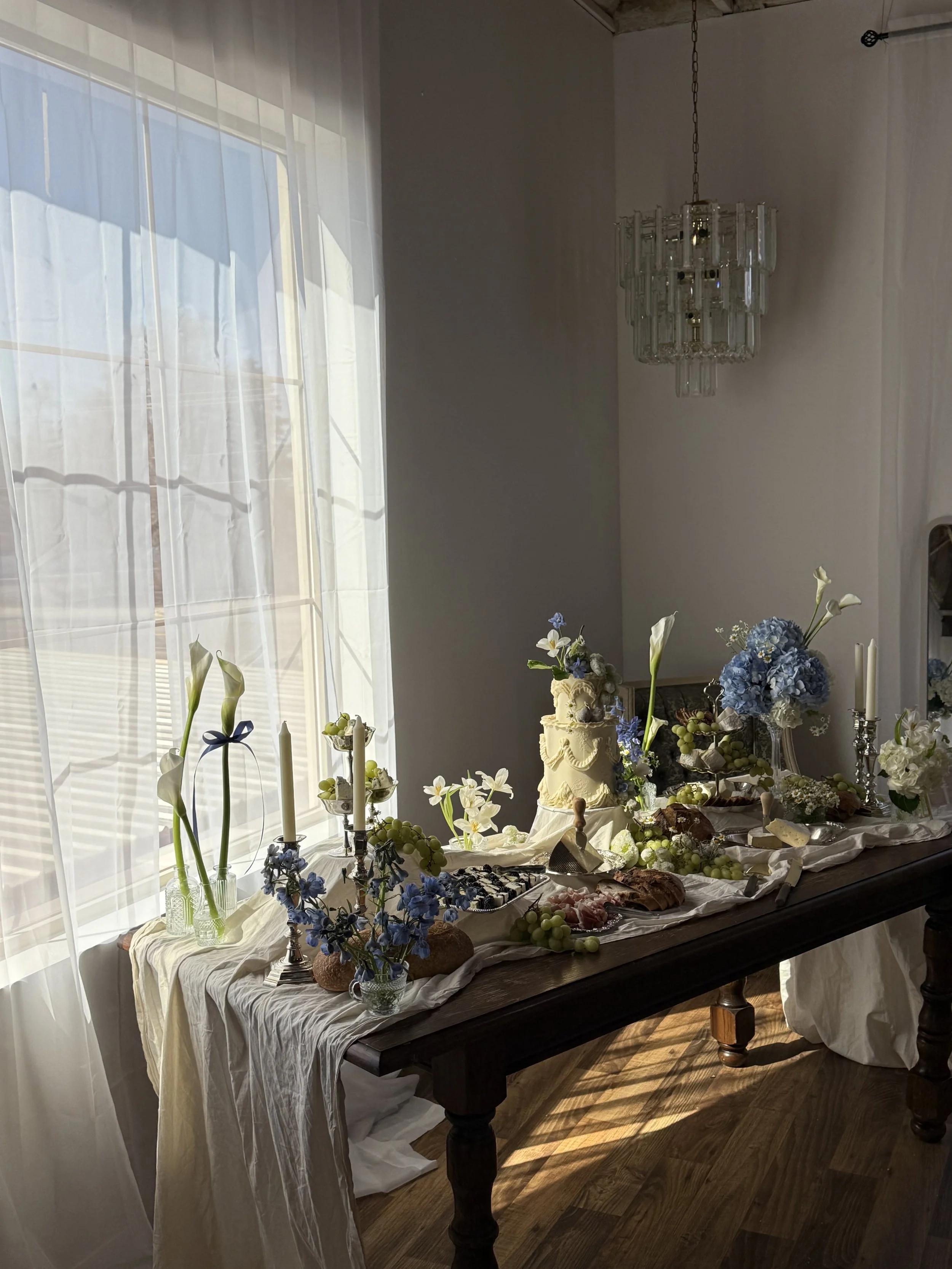 A dessert table set in a well-lit room with sheer curtains, decorated with flowers, candles, and a cake, featuring a vintage style with a crystal chandelier hanging above.