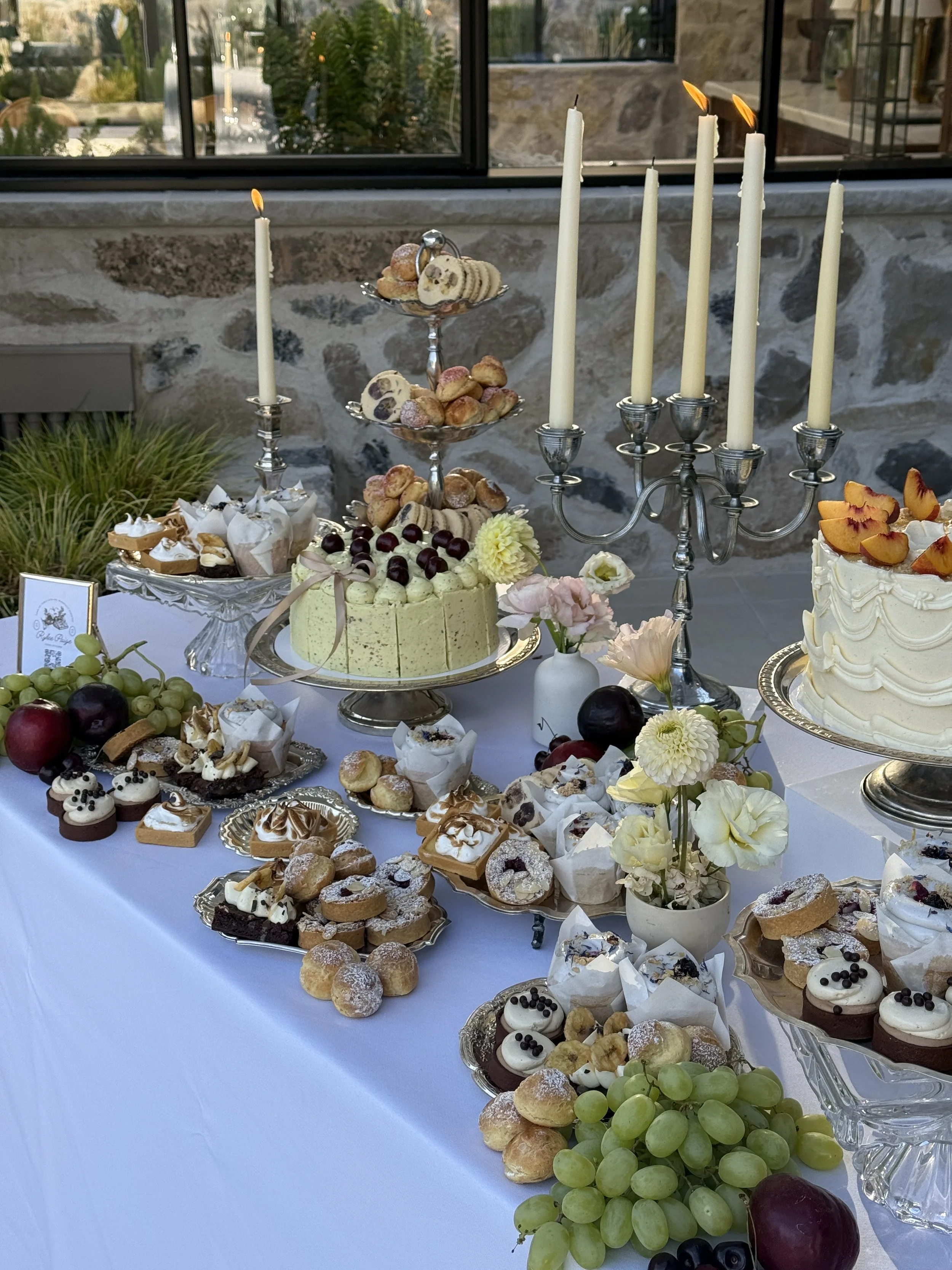 Table set with cakes, pastries, grapes, and a candelabrum at an outdoor celebration.