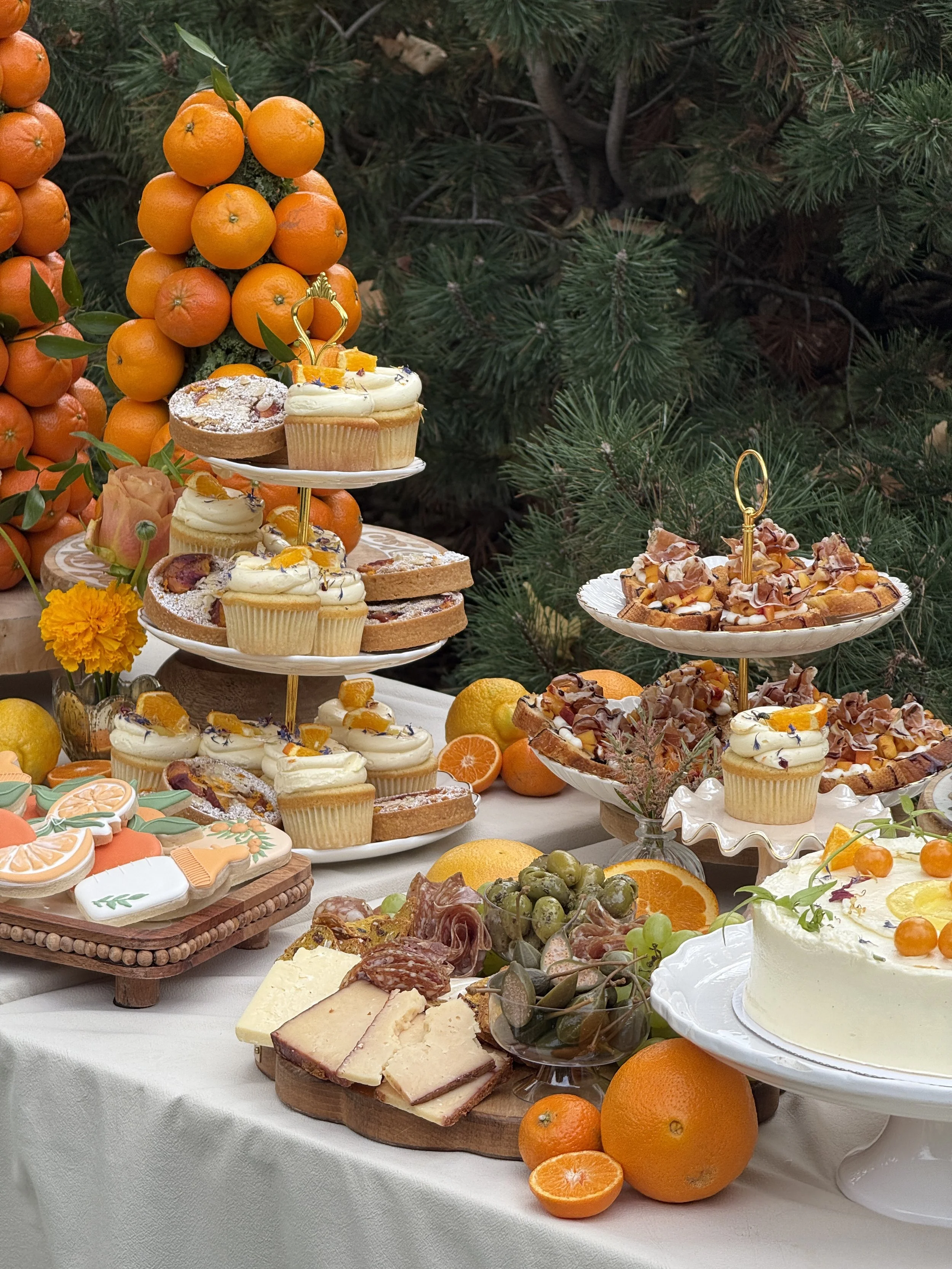 A dessert table featuring cupcakes, cookies, cake, and various fruits, set outdoors with greenery and small orange trees in the background.