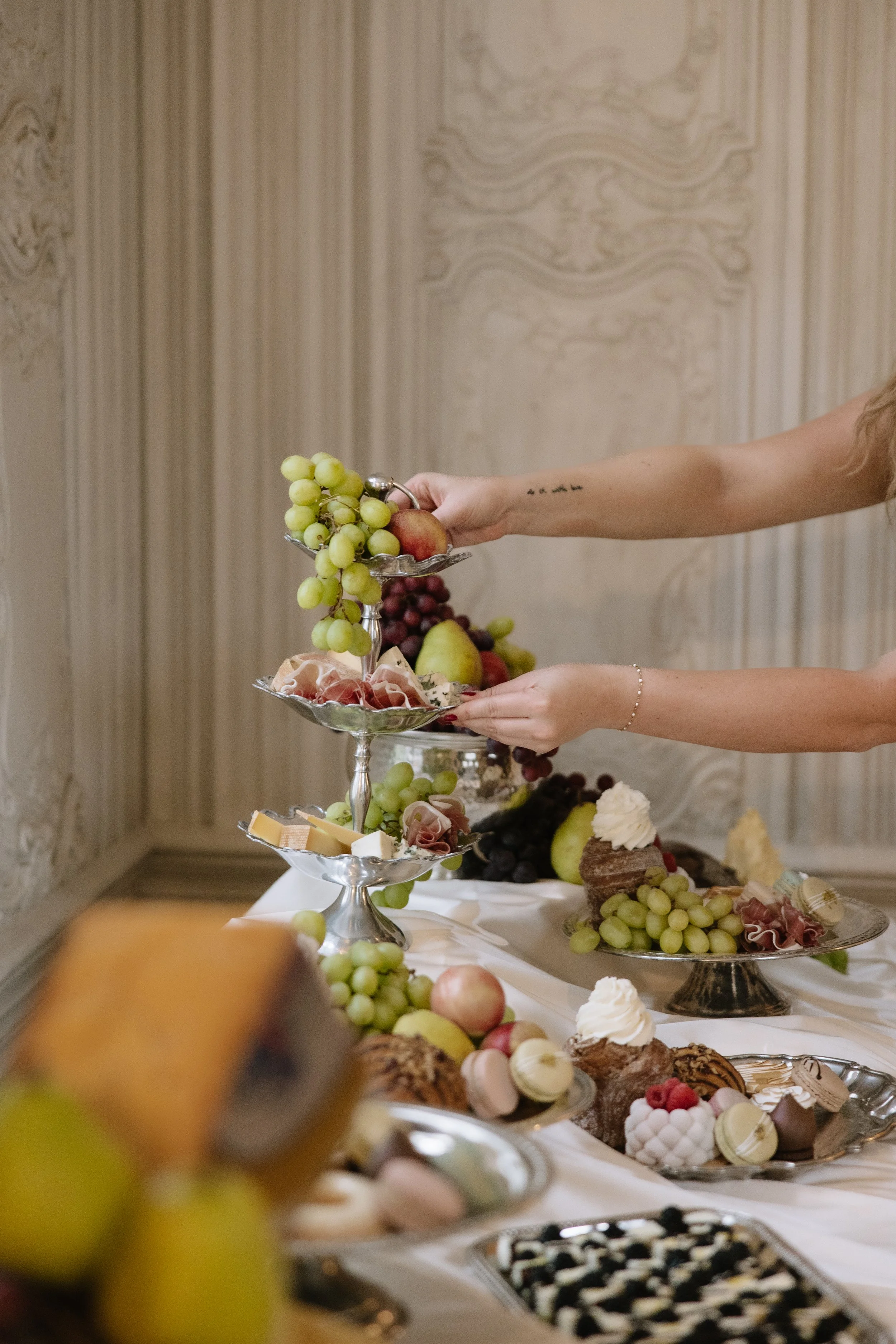 A person arranging a tiered tray of grapes, apples, and cheese on a buffet table with desserts and fruits in the background.