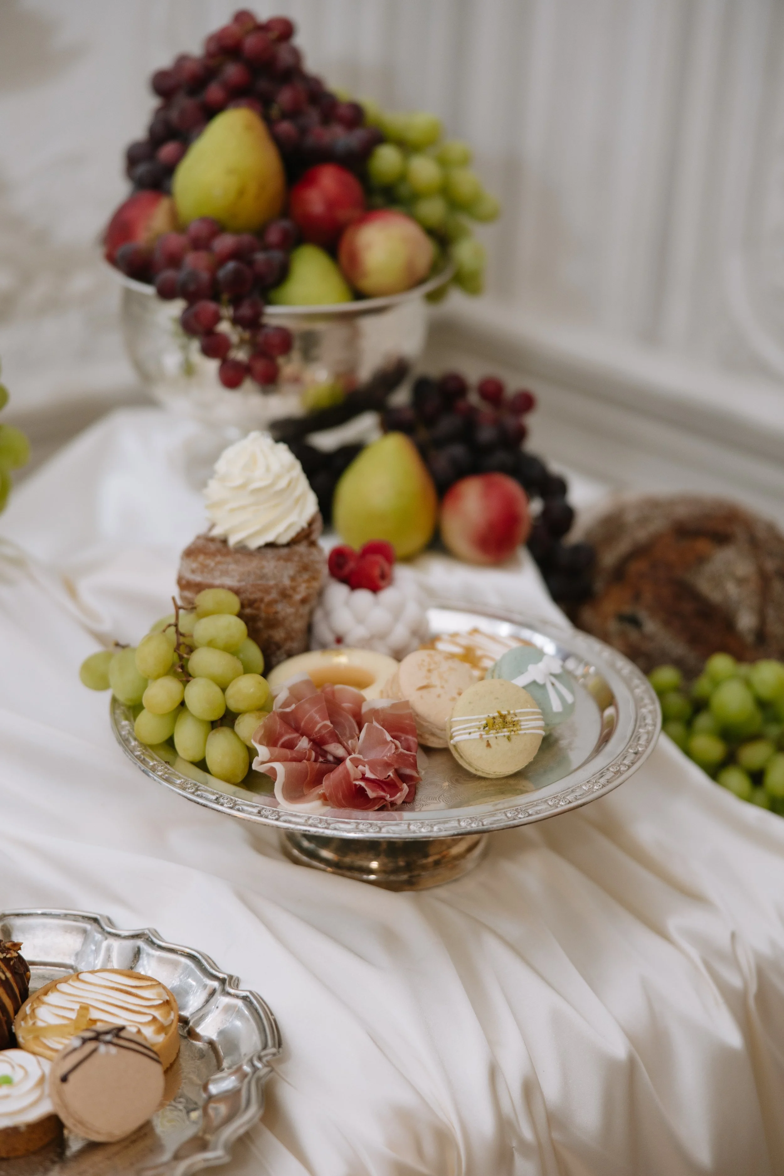 Assorted fruits and desserts arranged on a silver platter and glass bowls, including grapes, pears, apples, and macarons, on a white satin tablecloth.