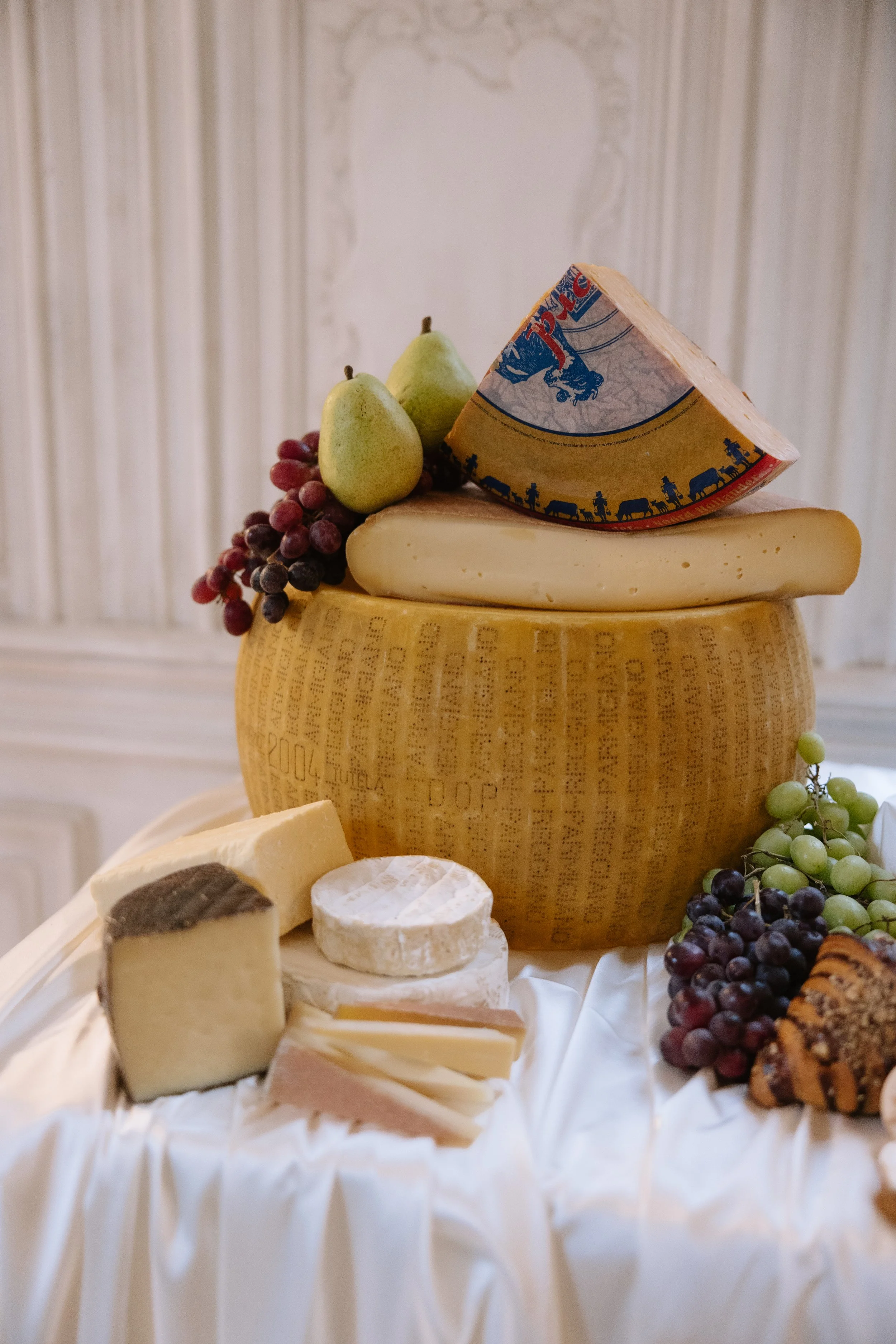 A cheese wheel topped with pears, grapes, and a slice of cheese, with various cheeses and grapes arranged on a white cloth beneath it.