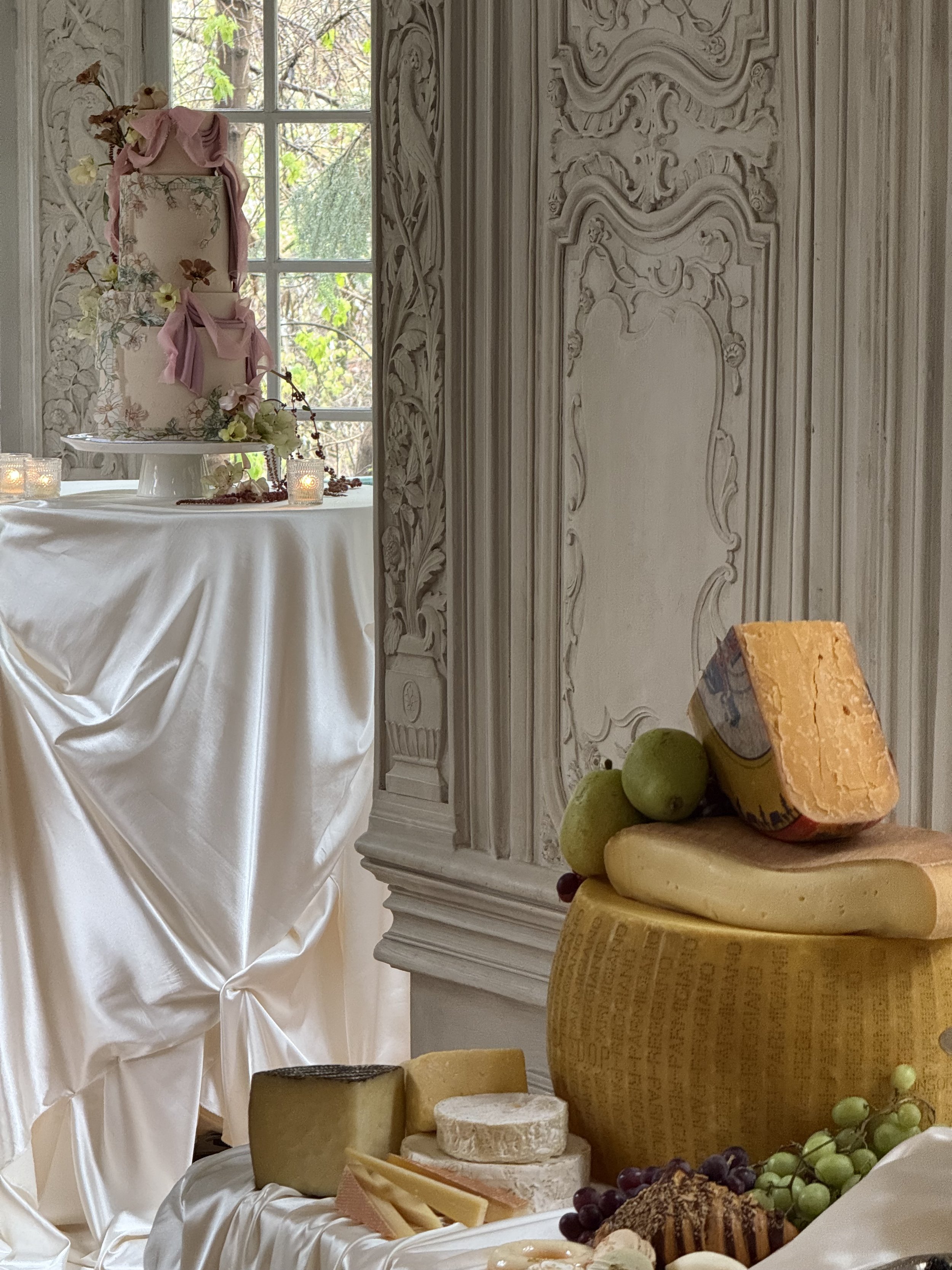 A decorated table with cheese, grapes, and slices of bread, with a large round cheese wheel in the background and a decorative cake on a stand nearby, set against an ornate wall and window in a bright room.