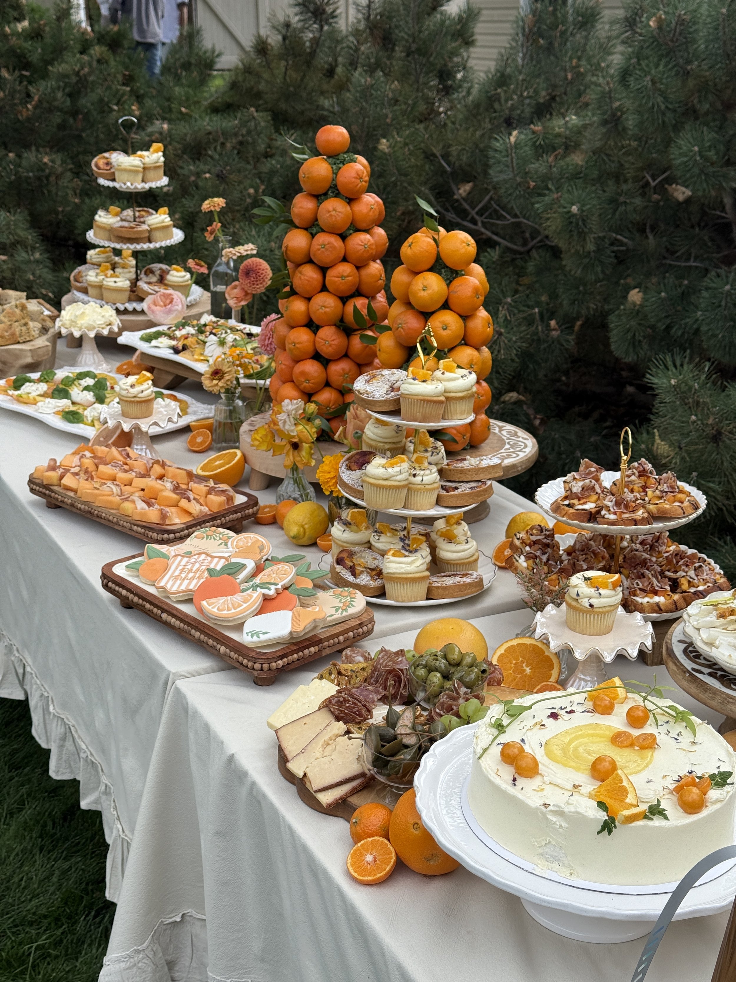 Outdoor dessert table with cakes, cupcakes, citrus fruits, and decorative oranges, set against greenery.