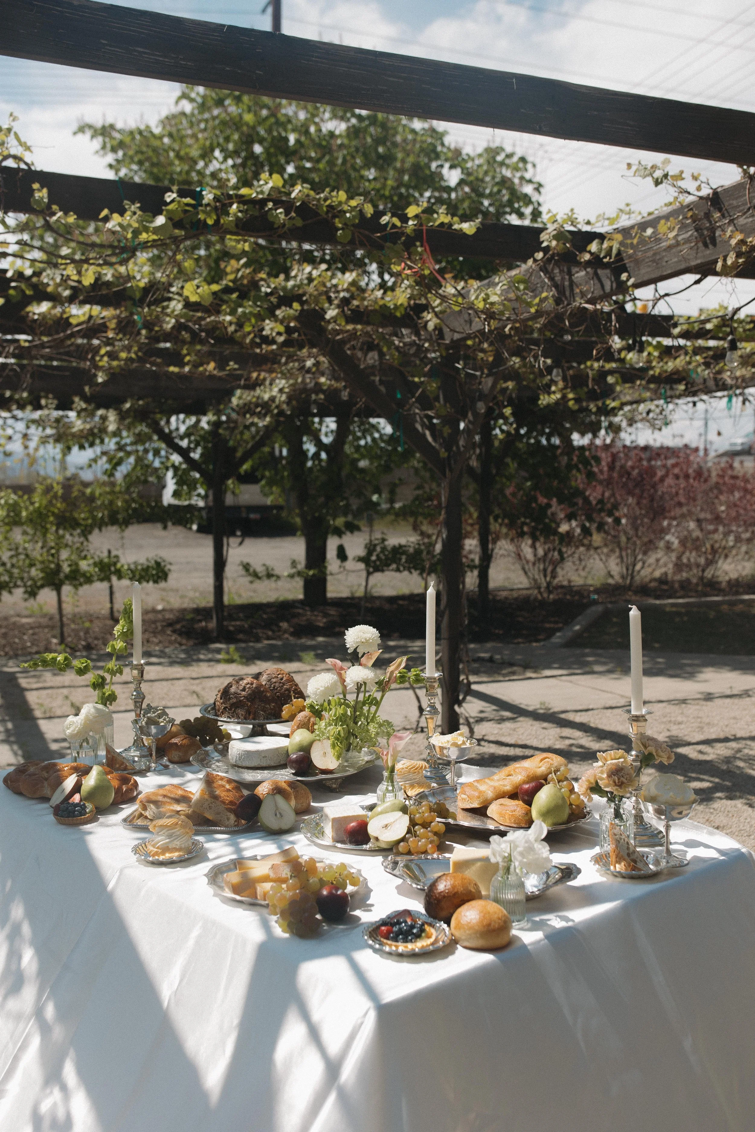 A table set with various foods including bread, fruits like grapes and pears, and desserts, in an outdoor setting under a pergola with plants, with trees in the background