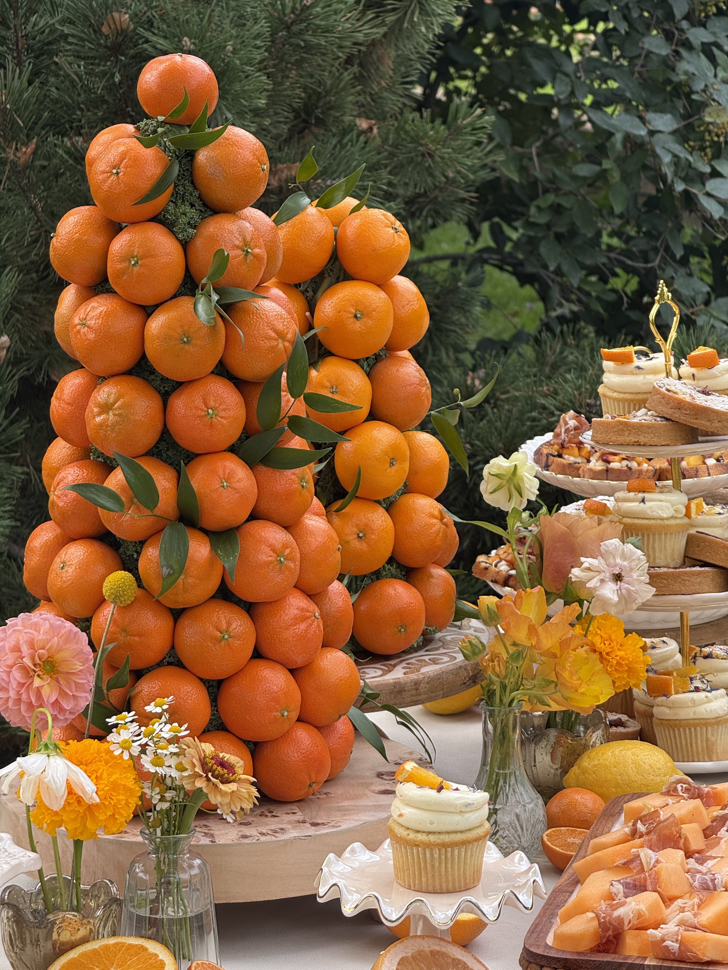 A large arrangement of stacked tangerines with green leaves, surrounded by flowers and a dessert table with cupcakes and fruit.