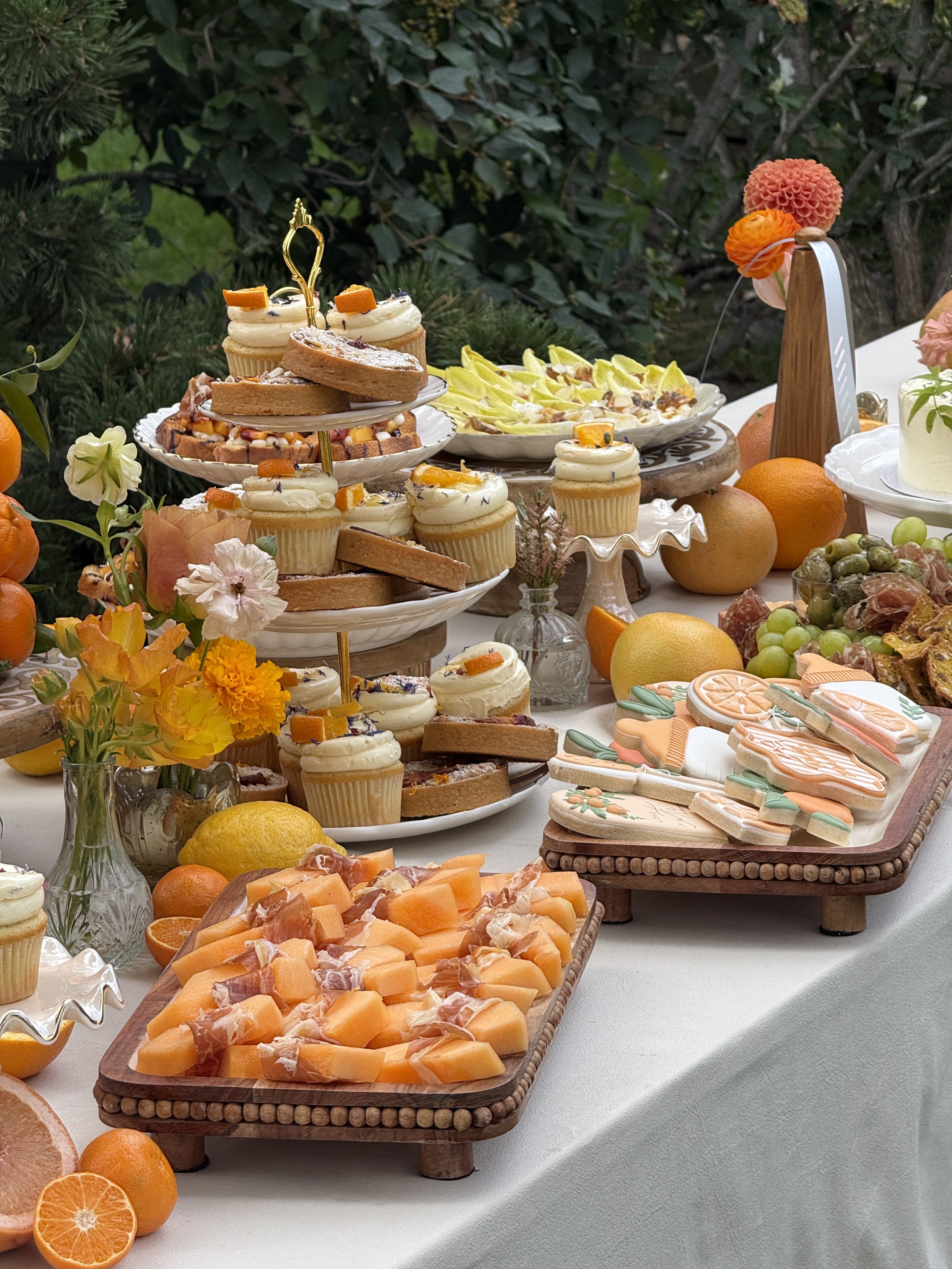 An outdoor dessert table with various sweets, including cupcakes, cookies, and finger sandwiches, surrounded by oranges, lemons, grapes, and flowers.