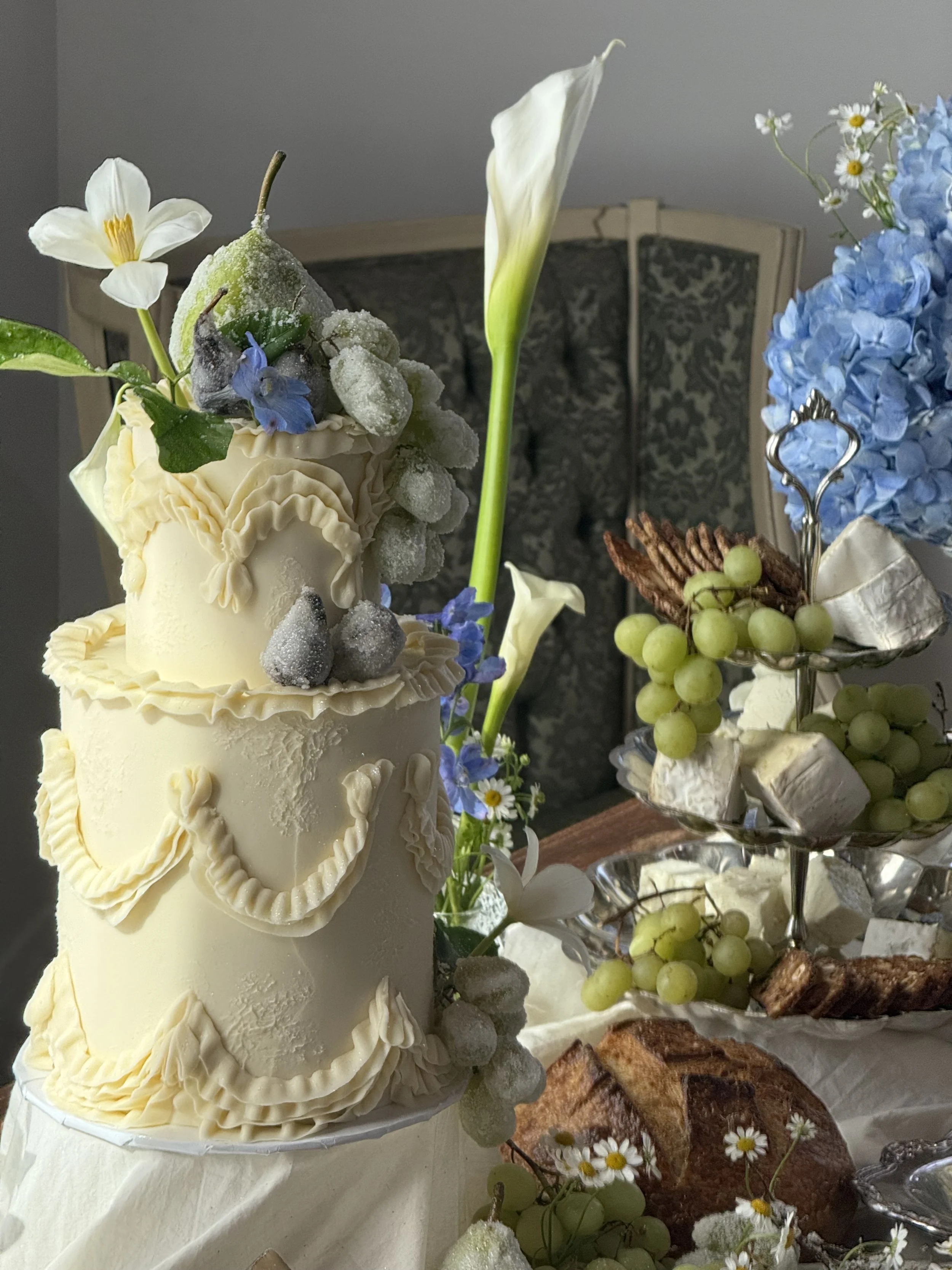 Elegant white tiered cake decorated with flowers and fruit, surrounded by flowers and baked goods.