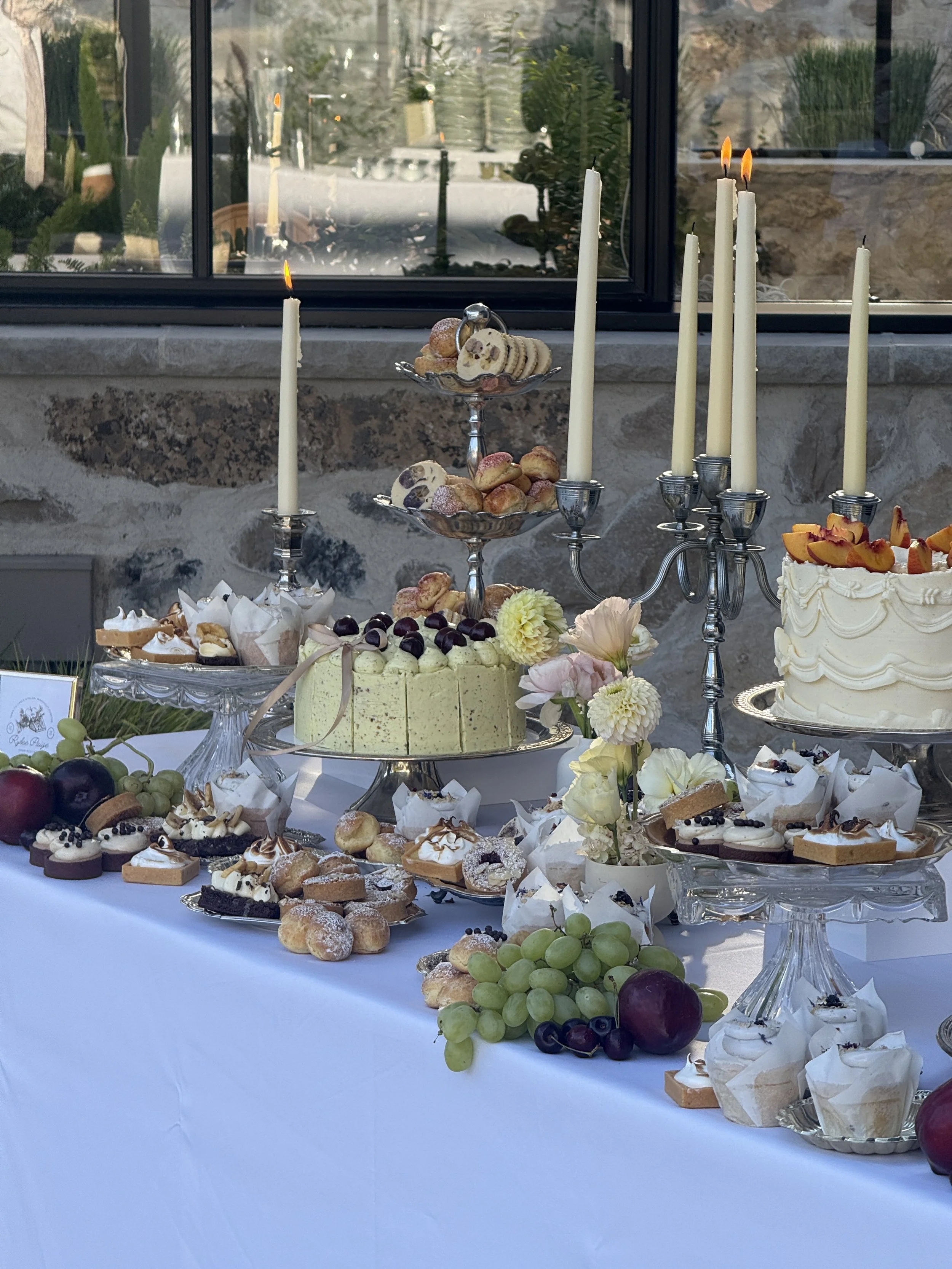 A dessert table with cakes, cookies, and fruit, featuring lit candles and floral decorations, set against a stone wall and window.