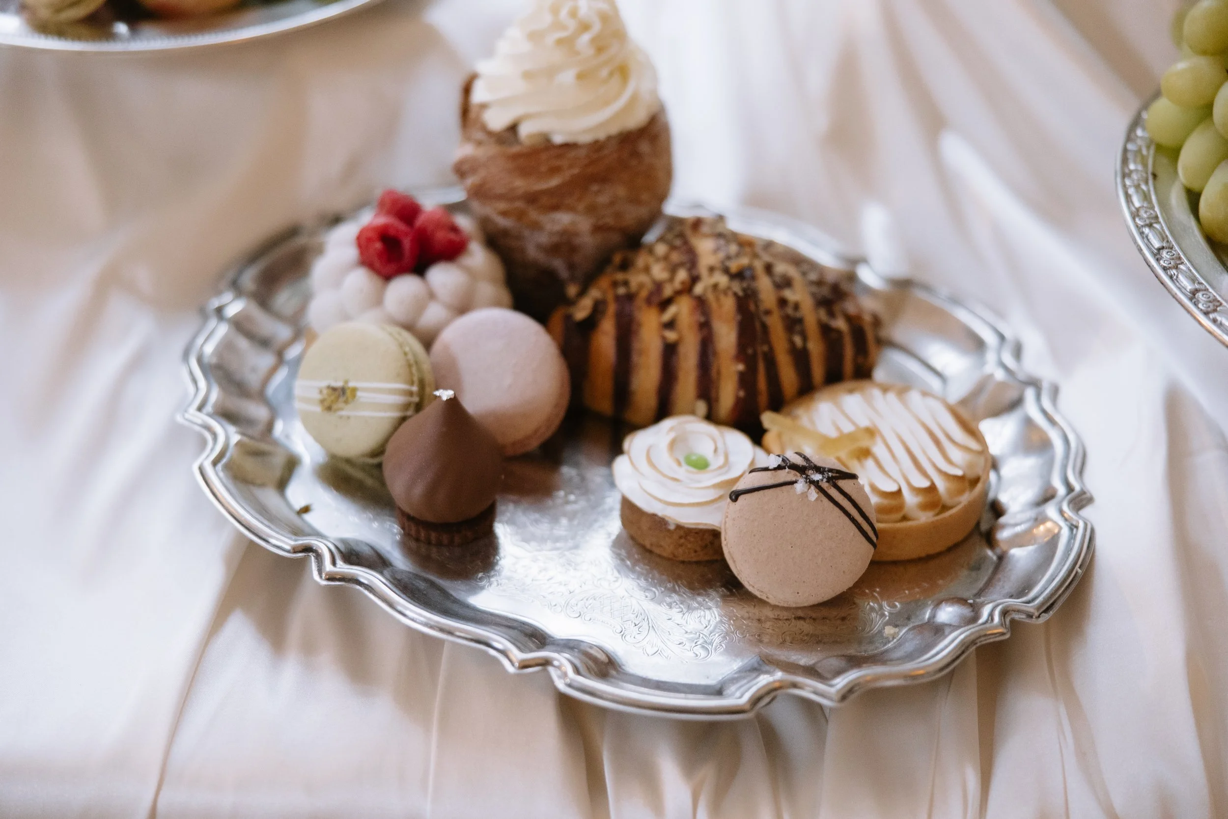 Assorted pastries and desserts on a silver platter, including macarons, tarts, and cream-filled baked goods, arranged on a white tablecloth.
