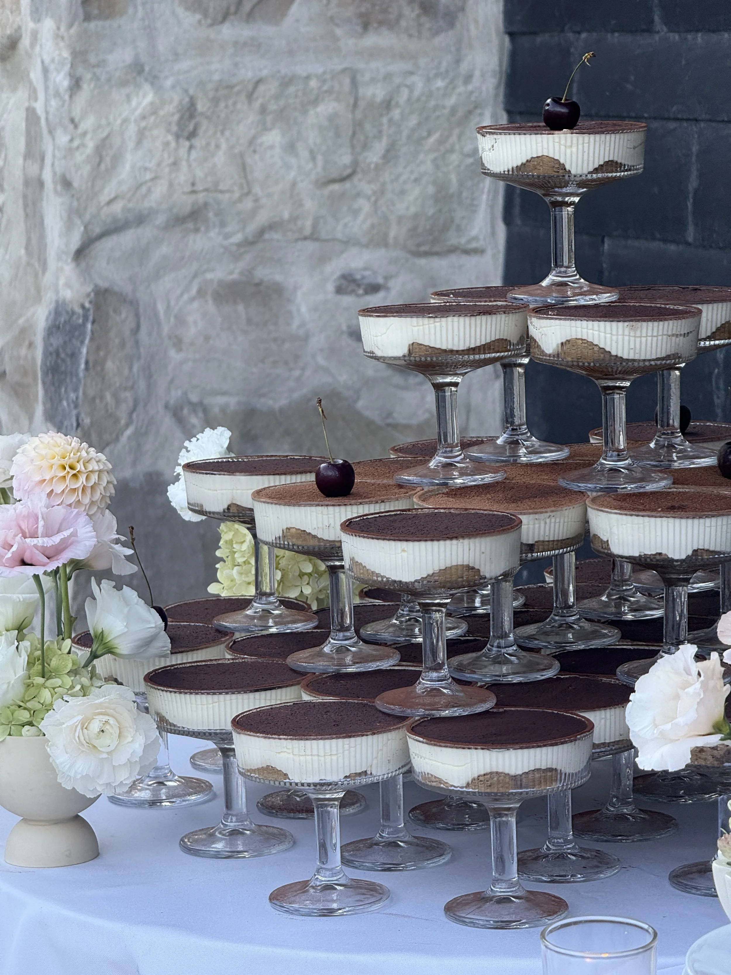 A tiered display of individual tiramisu desserts in glass cups, decorated with cocoa powder, with cherries on top, set on a table with flowers around.