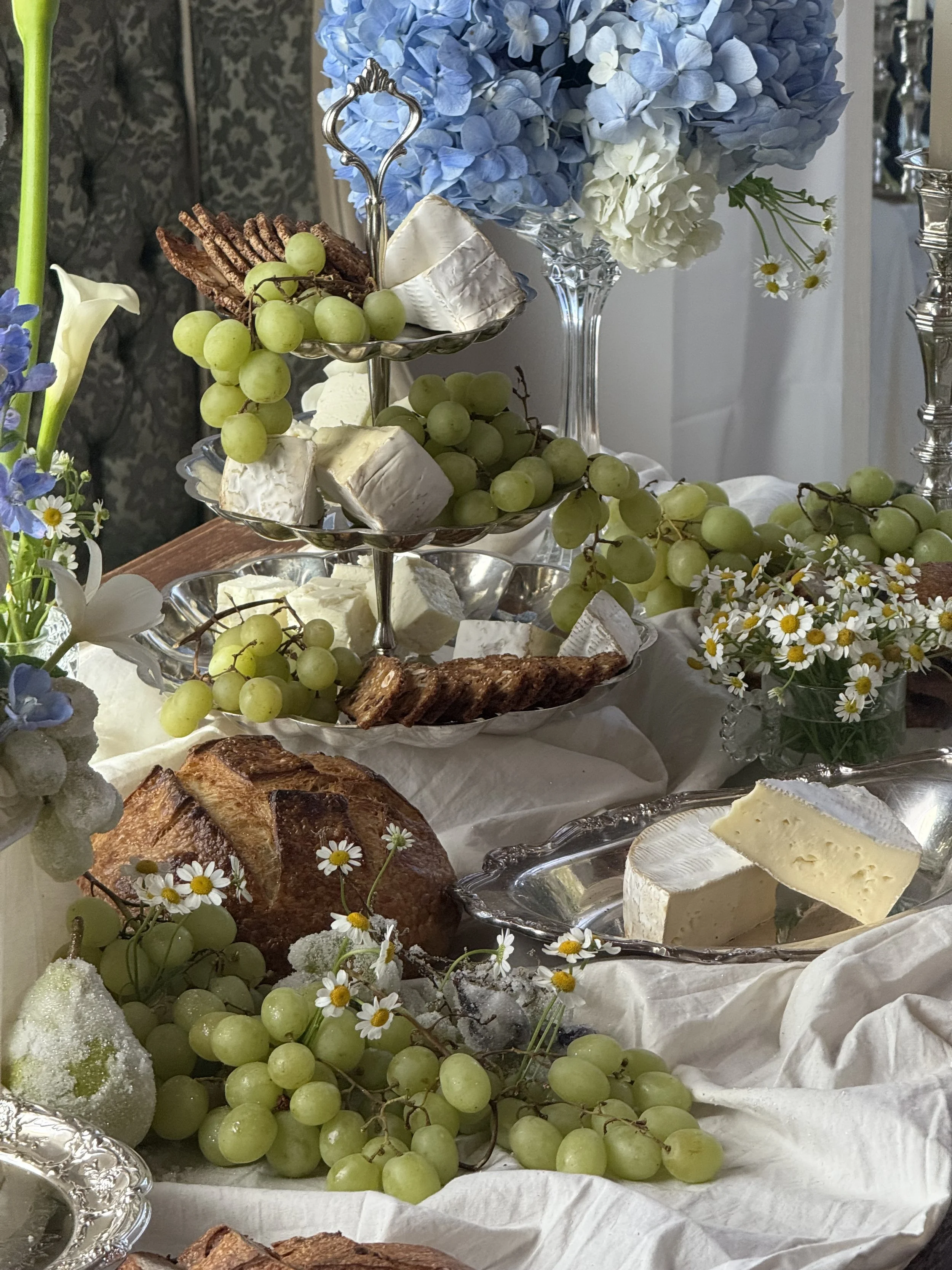 A cheese and fruit platter with grapes, cheeses, baked bread, and floral decorations on a table.