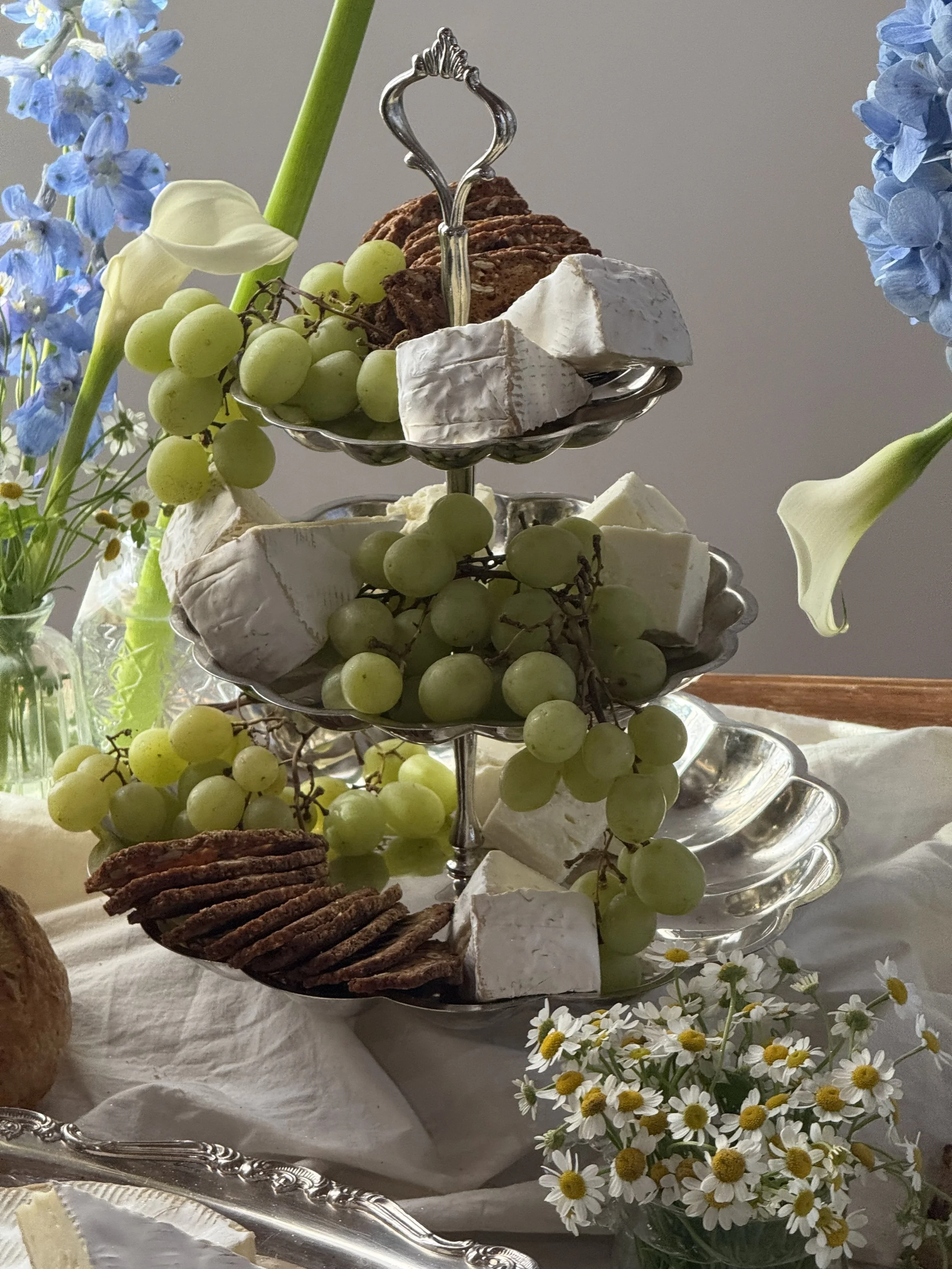A three-tiered silver serving tray holding grapes, Brie cheese, chocolate cookies, and chocolate cake, surrounded by daisies and blue hydrangeas on a white tablecloth.
