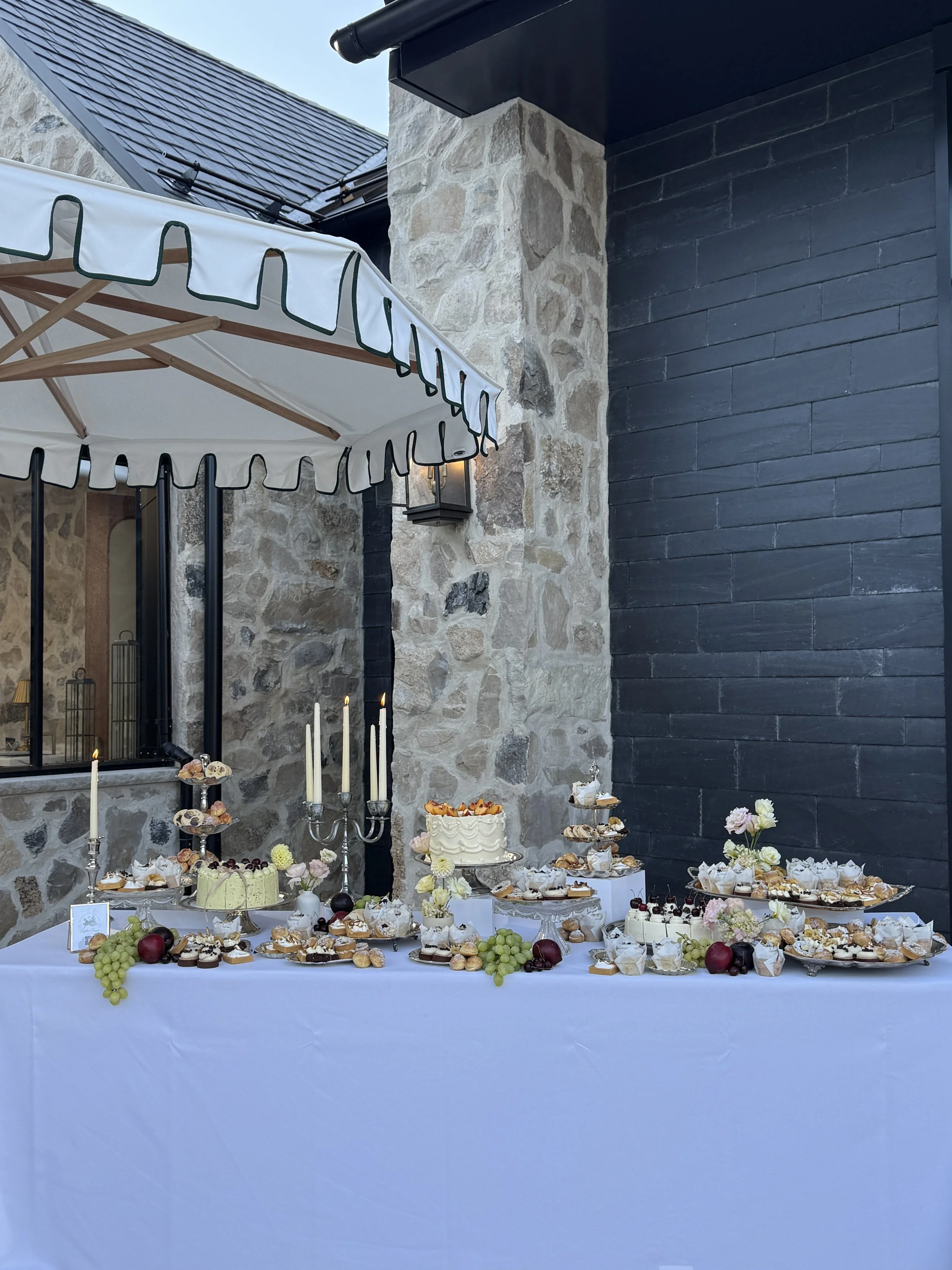 Dessert table with cakes, pastries, and fruits, decorated with flowers and candles, set outdoors under an umbrella with stone and black wall background.